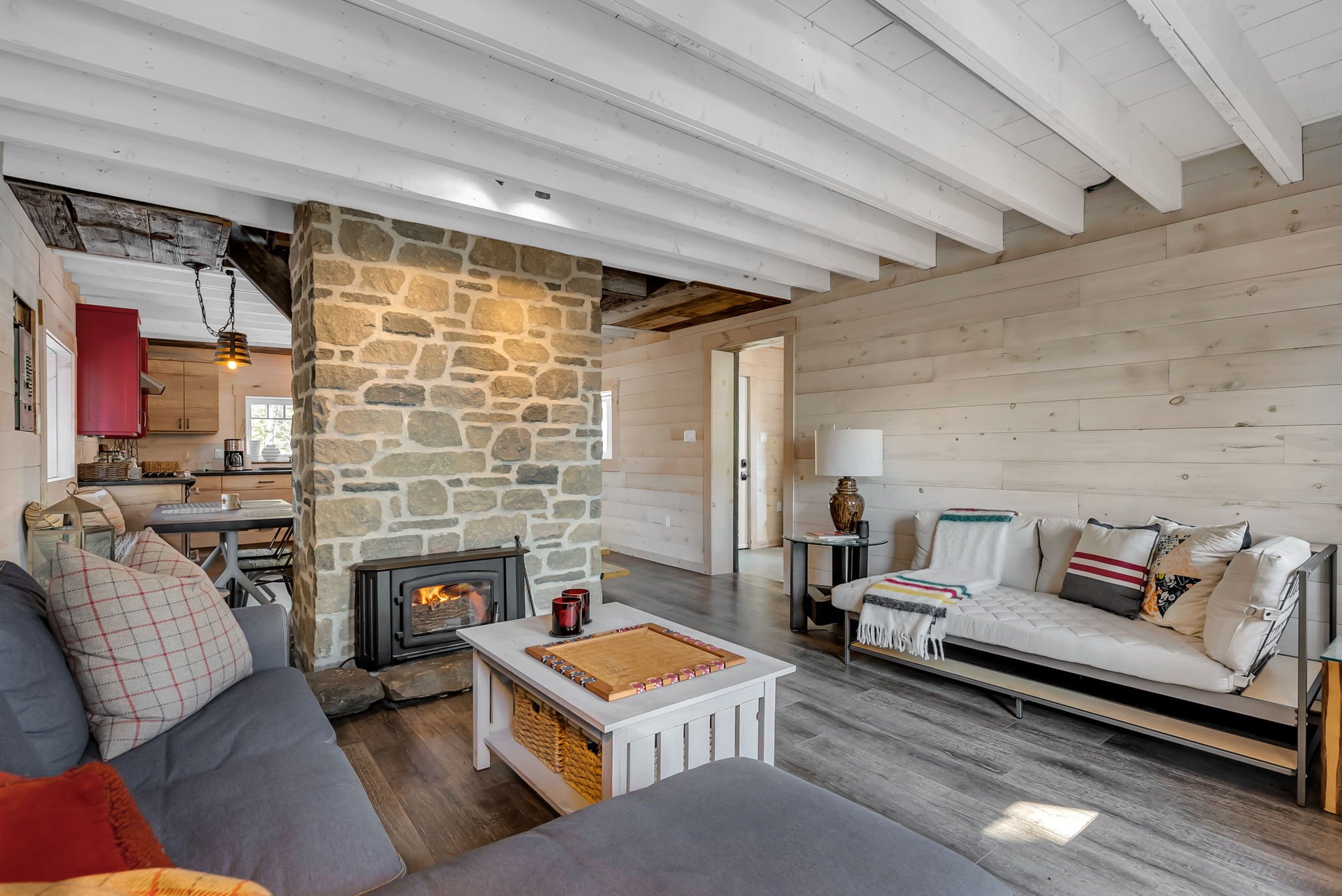 Open-concept living space in an Ottawa home featuring a gray sofa, wood stove with stone chimney, and white wood-paneled walls, with a connected kitchen and island seating in the background, creating a warm and functional layout.