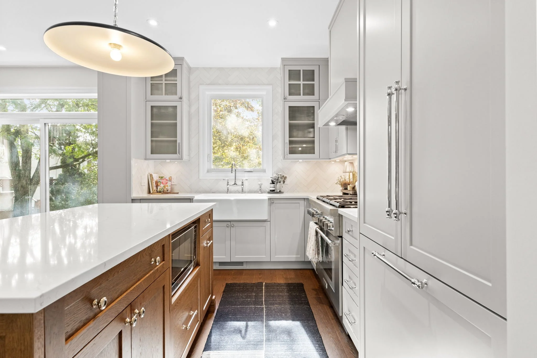 Kitchen with white cabinets, a white farmhouse sink under a window, a black rug on wooden floor, and a modern pendant light fixture.