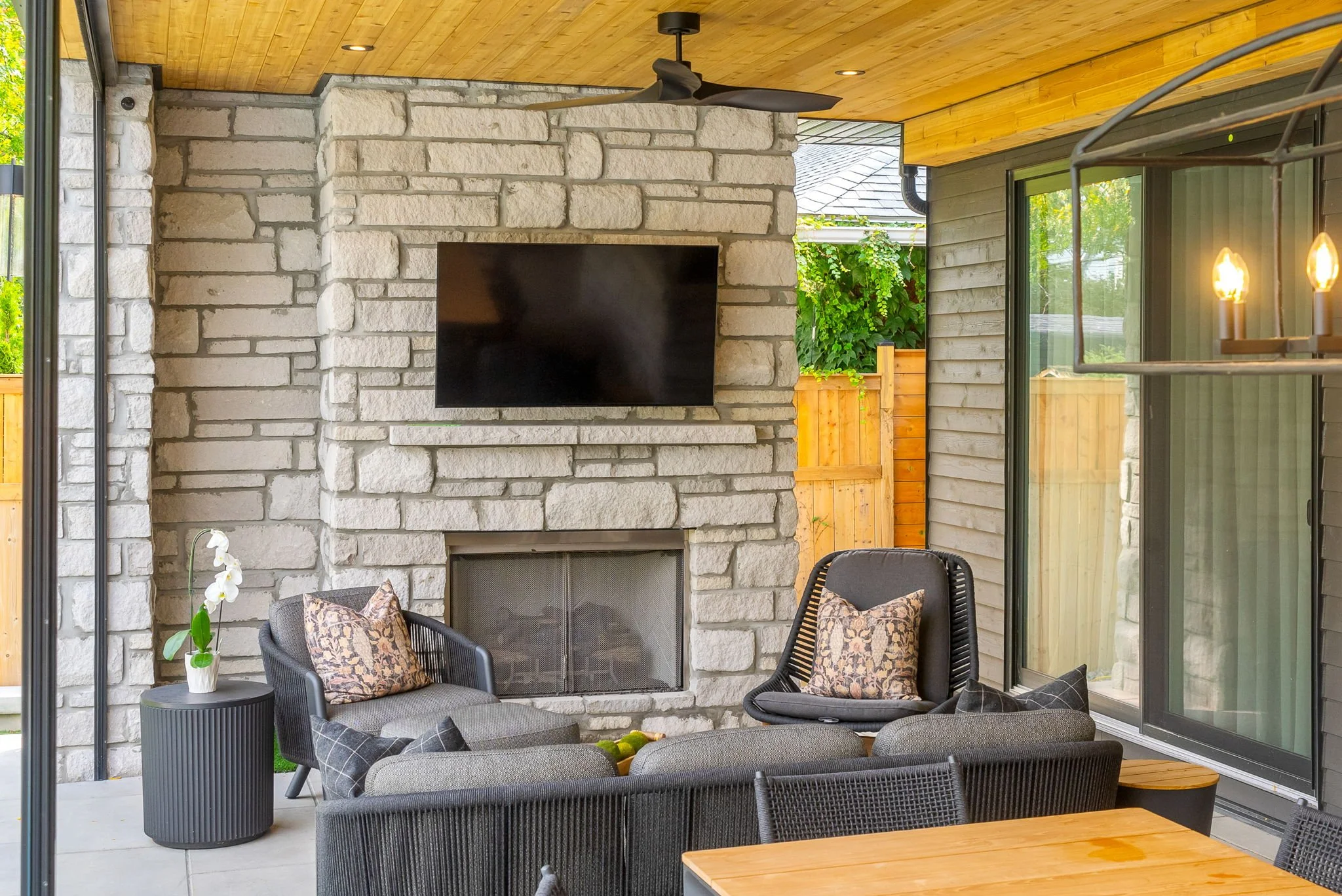 Outdoor living space with a stone fireplace, mounted flat-screen TV, ceiling fan, sectional sofa with cushions, side table with a vase of white flowers, and a wooden table, enclosed by a wooden and glass structure.