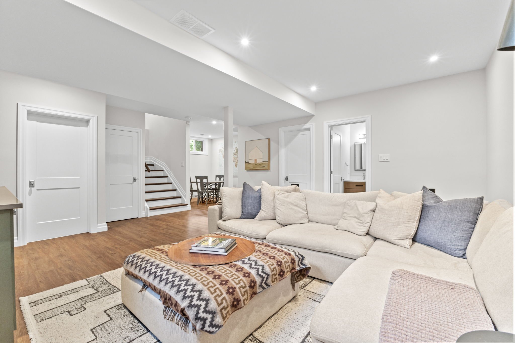 Open-concept living room in an Ottawa home featuring a beige sectional, hardwood flooring, and recessed lighting, flowing into a dining area and staircase, designed for comfortable everyday living and seamless layout