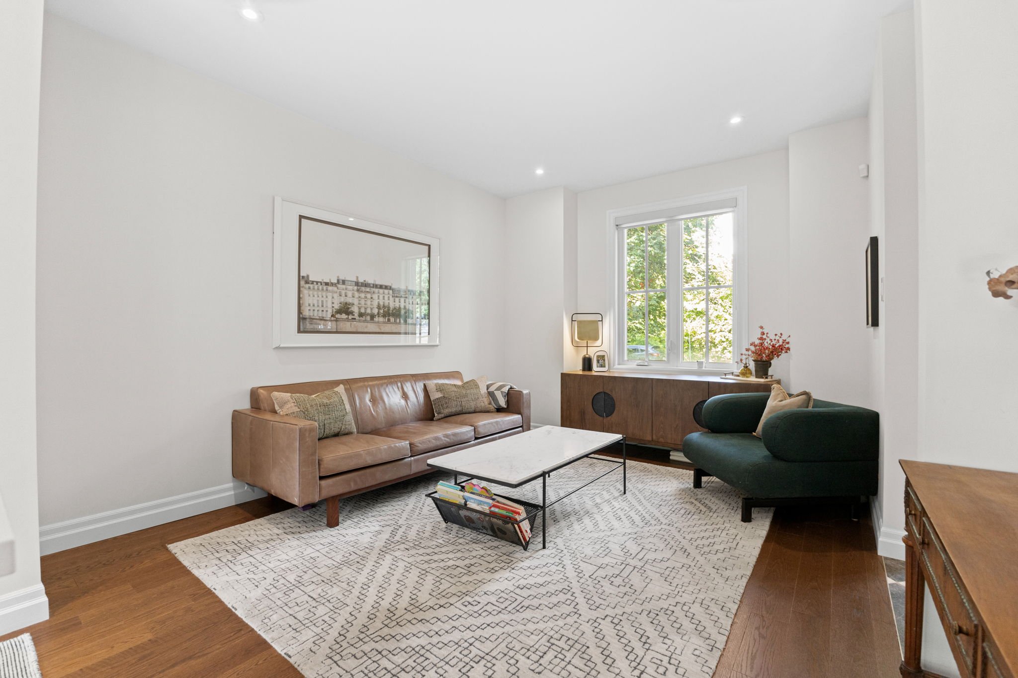 Living room with a tan leather sofa, a dark green armchair, a white coffee table, patterned rug, wooden sideboard, and window with greenery outside.