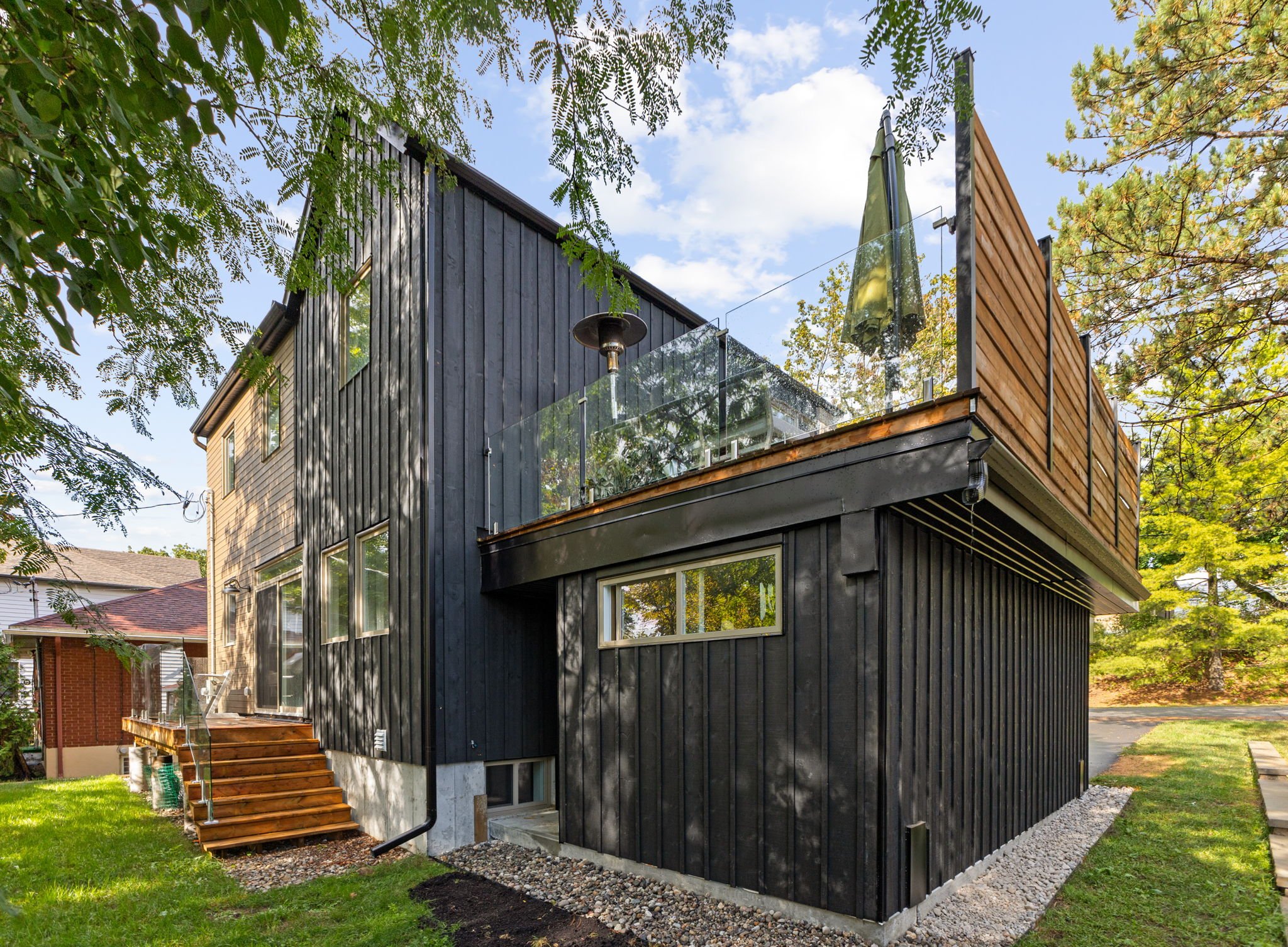 A modern two-story house with black and wooden exterior panels, large windows, a small staircase leading to a back door, and a rooftop terrace with a glass railing and a patio umbrella, surrounded by trees and a lawn.