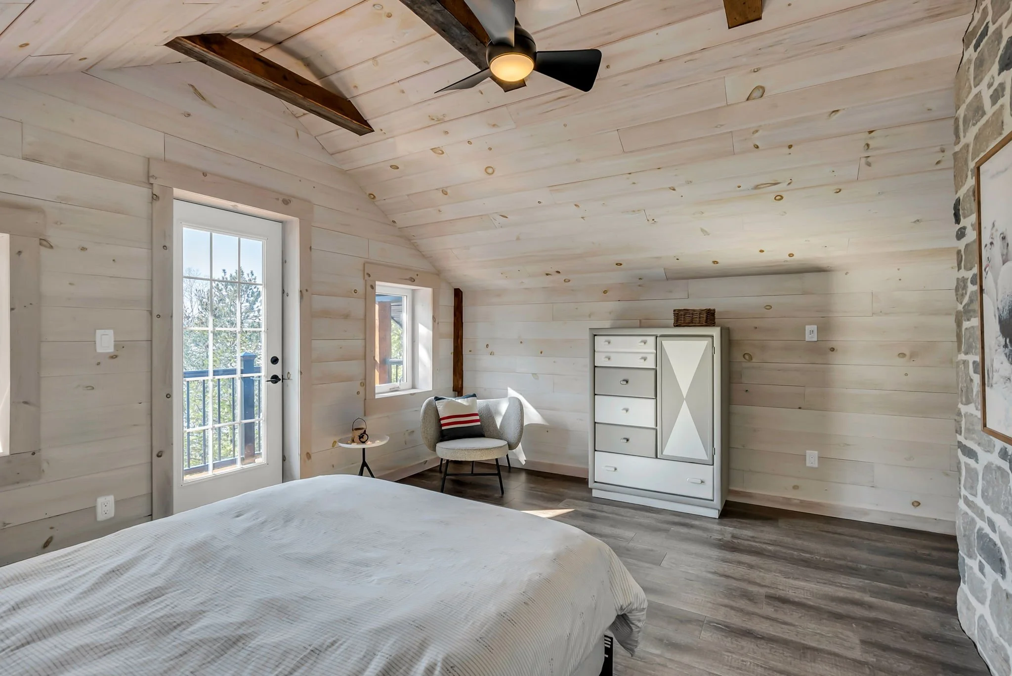 Cozy bedroom in an Ontario home featuring wood-paneled walls, a comfortable bed, white dresser, and seating area, with a sliding glass door providing direct outdoor access and natural light, designed by Centennial Construction Management.