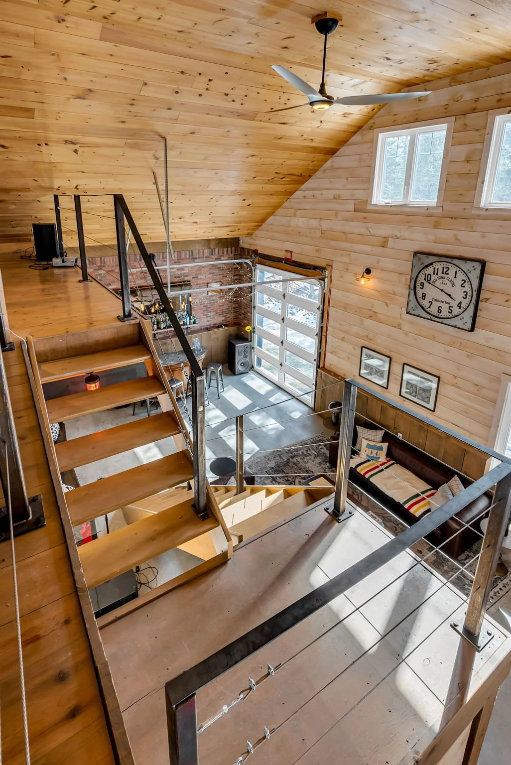 Interior of a modern, rustic-style home with wooden walls and ceiling, featuring stairs with metal railing, a loft area, large windows, a garage door, a clock, and cozy furniture in the living space.