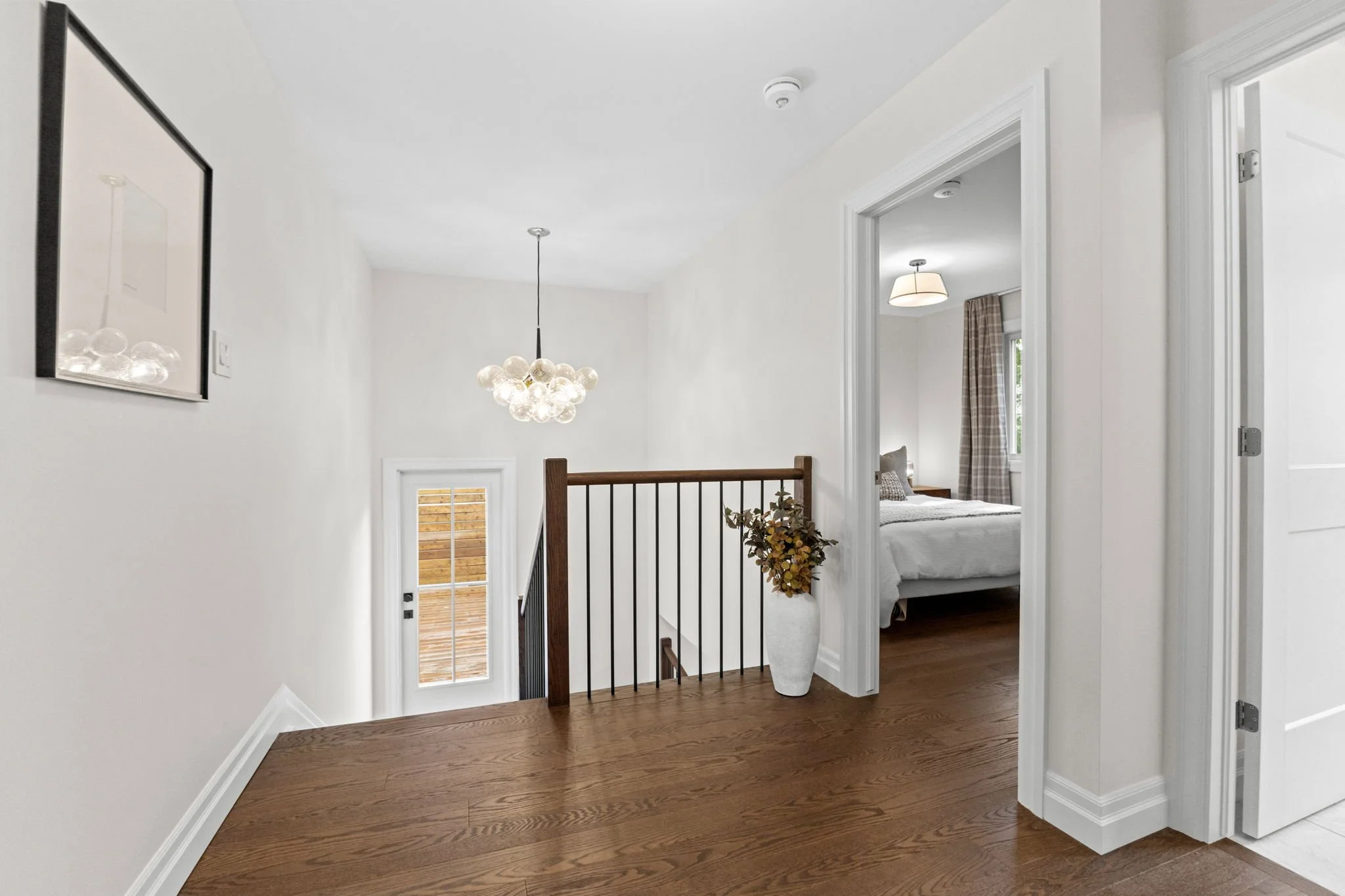 Second-floor hallway in a modern Ottawa home featuring hardwood flooring, a wood-railed staircase, and a statement glass globe chandelier, with sightlines into a bright bedroom, showcasing clean design and functional layout.