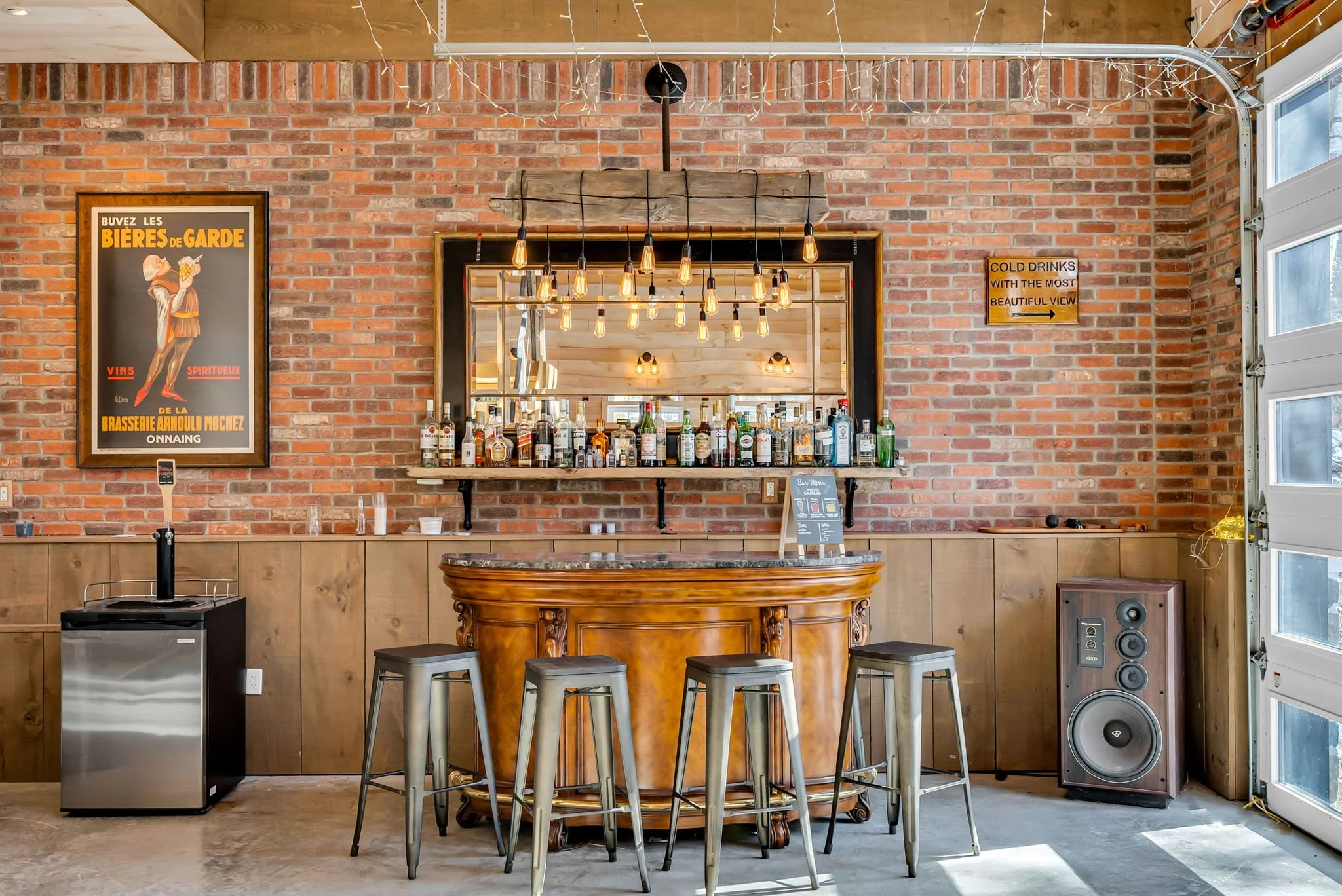 A cozy home bar with a brick wall background, a wooden bar counter, bar stools, a liquor shelf, a large mirror, a poster on the wall, and a speaker on the floor near a garage door.