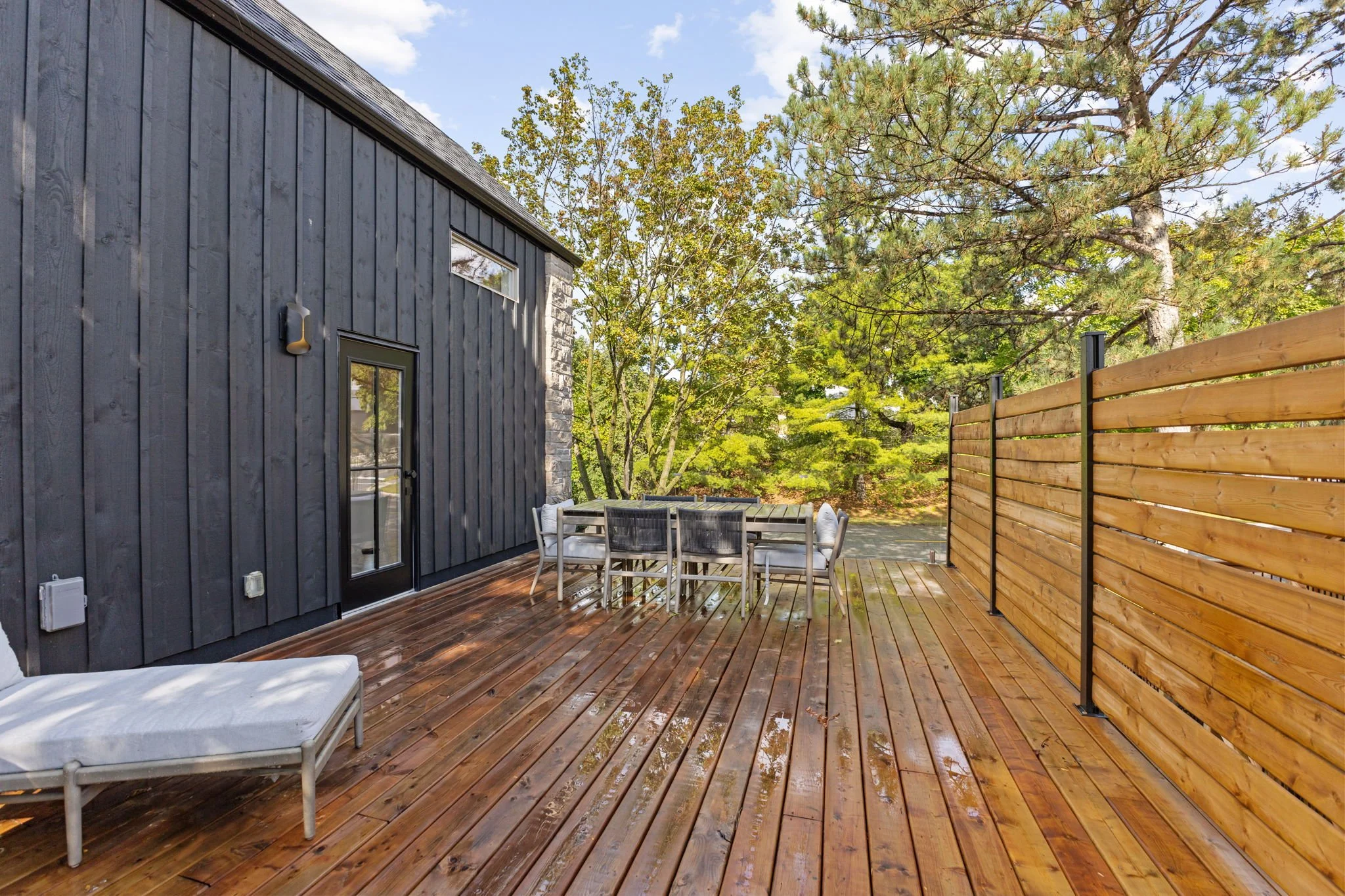 Wooden deck with outdoor dining table and chairs, a lounge chair, black vertical siding, a wooden fence, and trees in the background under a partly cloudy sky.
