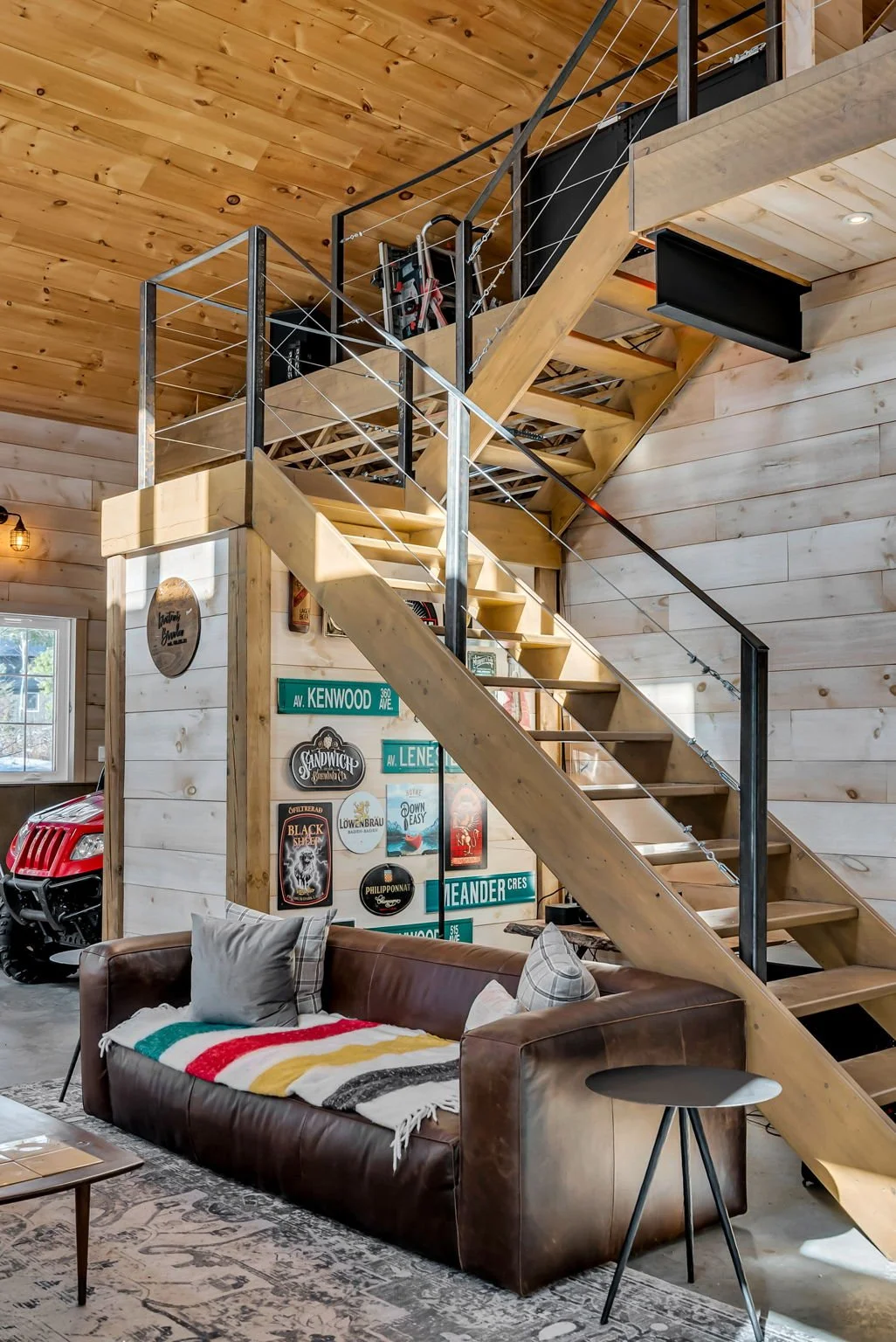 Interior of a home with a wooden staircase leading to an upper level, walls decorated with vintage signs, a leather couch with pillows and a blanket, a side table, and a vehicle partially visible in the garage area.