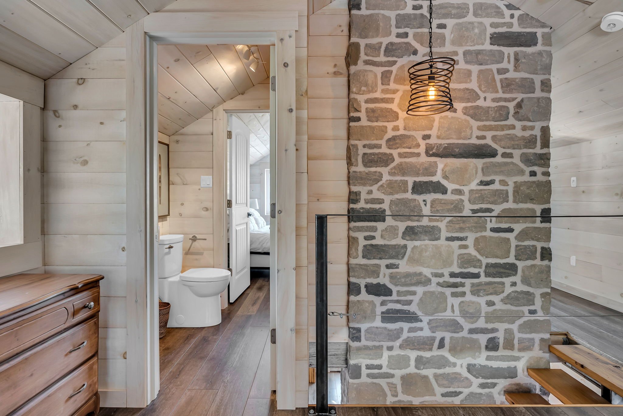 Interior view of a rustic-style house with wood-paneled walls and ceiling, featuring a stone wall with a hanging black spiral pendant light, a staircase with wooden steps, and a view into a bathroom and bedroom.