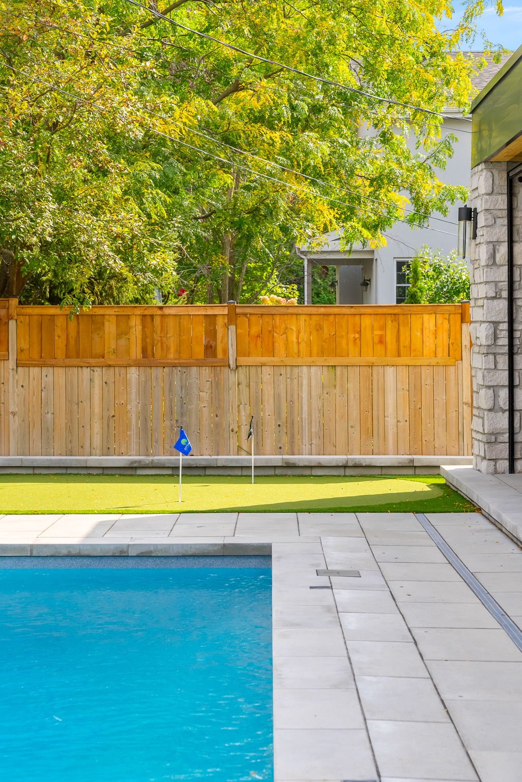 A backyard scene with a swimming pool, a green putting green with two small golf flags, a wooden fence, trees with green and yellow leaves, and part of a house with stone siding.