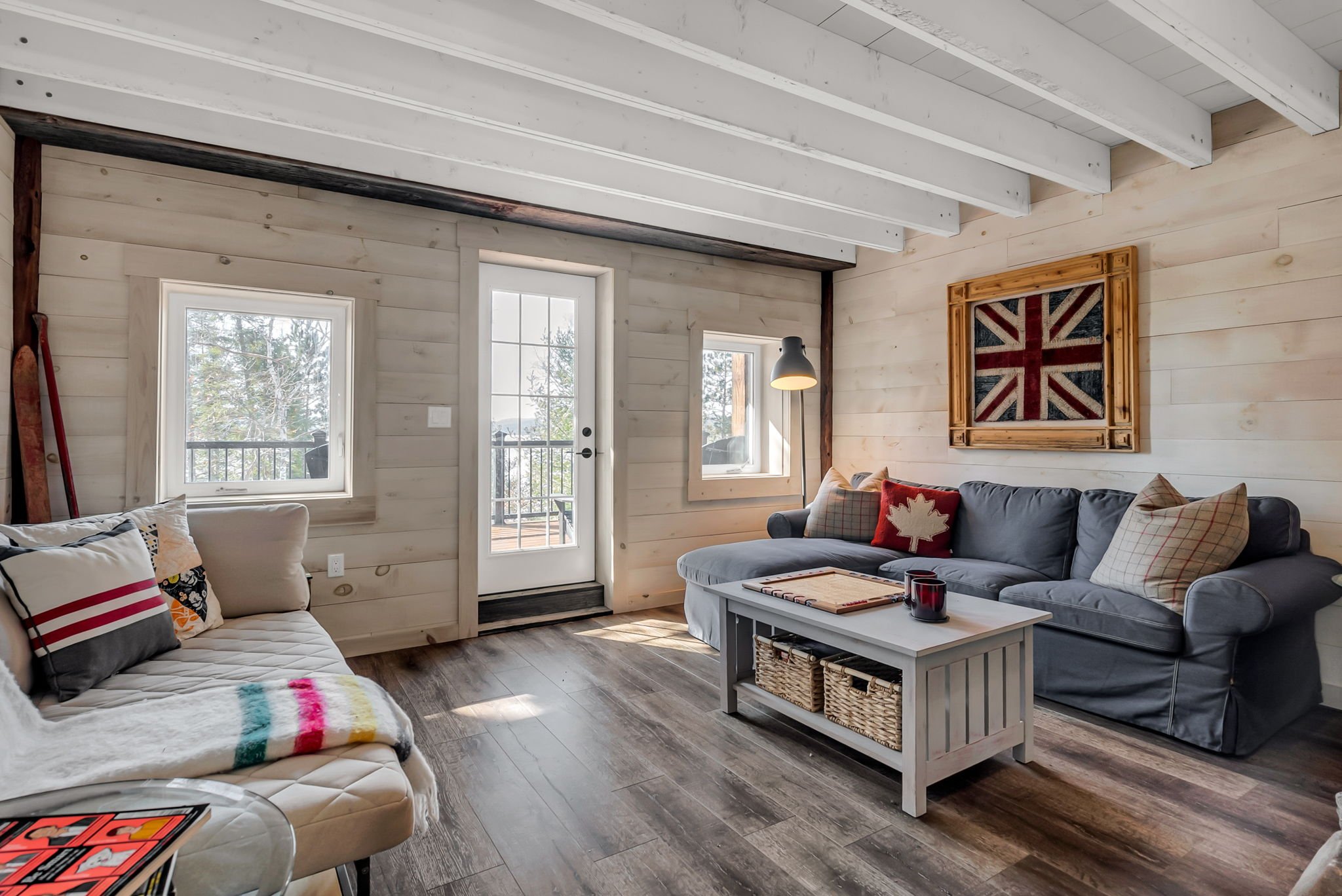 Cozy living room in a Rideau Lakes home featuring wood-paneled walls, white ceiling beams, and wide plank flooring, with layered seating, natural light, and curated décor creating a warm, cabin-inspired atmosphere.