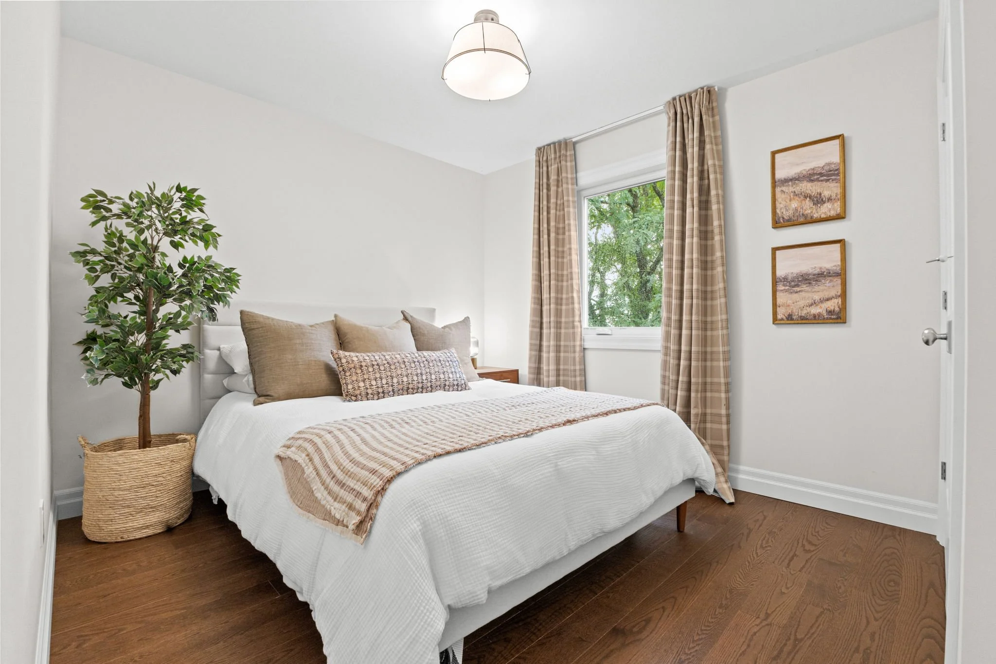 A cozy bedroom with a white bed, beige pillows, a throw blanket, a window with plaid curtains, a potted plant, and framed artwork on the wall.