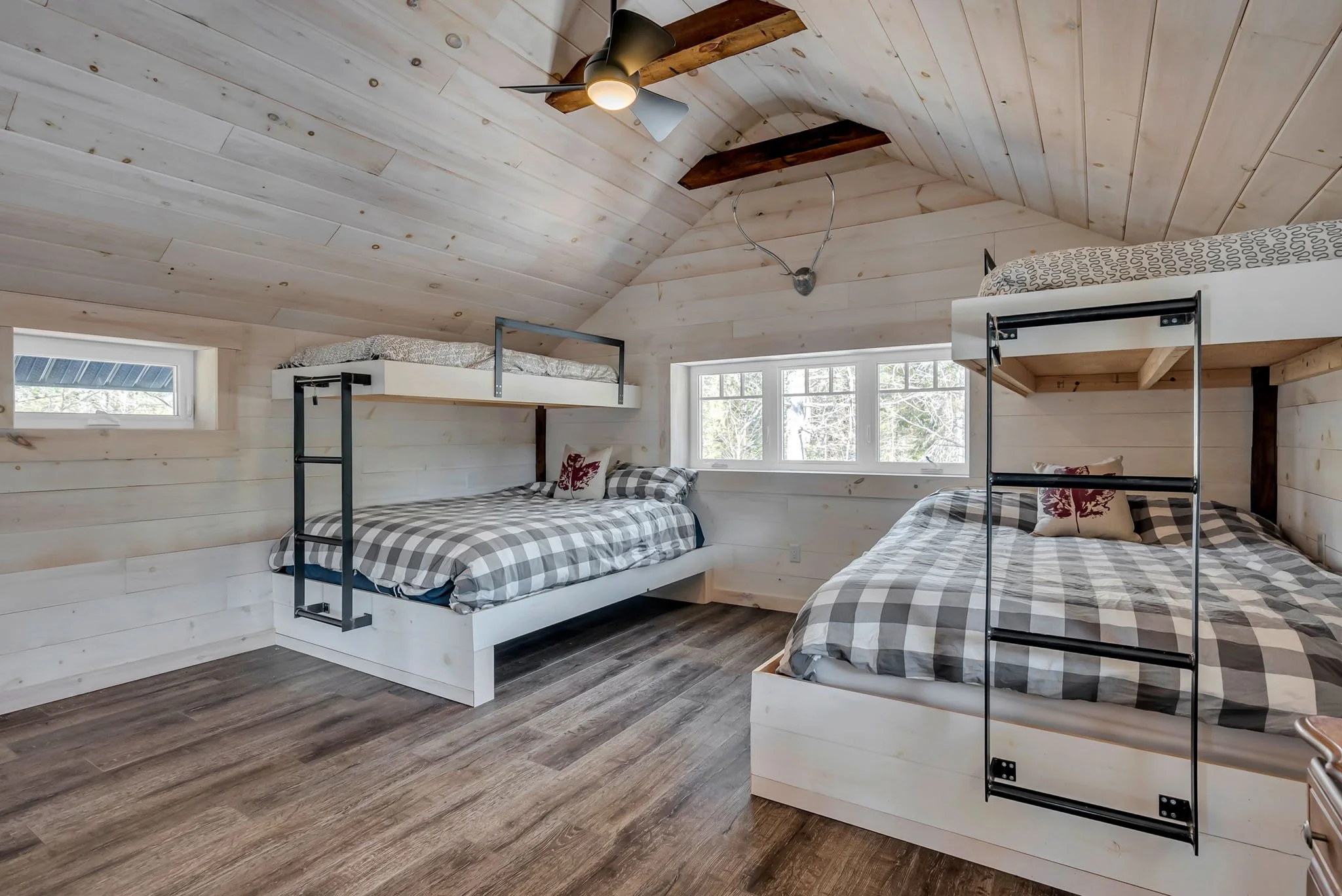 Custom bunk bedroom in an Ontario home featuring light wood-paneled walls and ceiling, built-in double beds with black metal ladders, and a ceiling fan, designed to maximize sleeping capacity with a warm, cabin-inspired aesthetic 