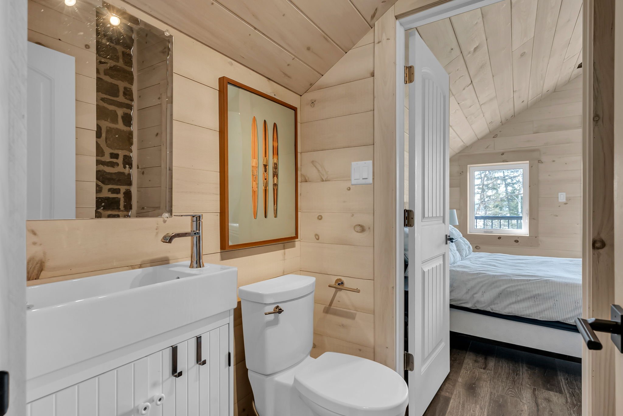 Bathroom with wooden walls, small white sink, framed ski artwork, white toilet, and a door leading to a bedroom with a bed, window, and wood-paneled ceiling.