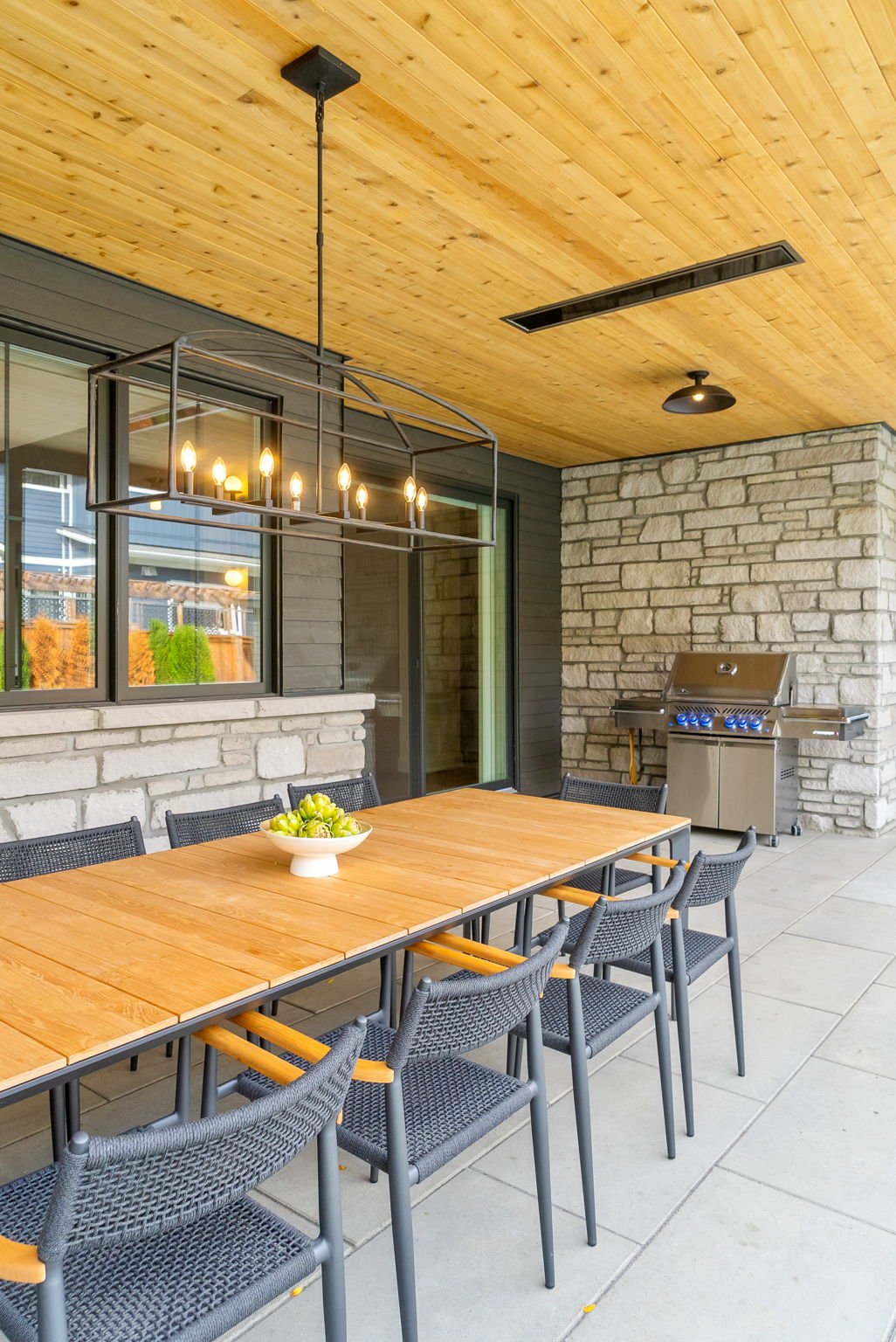 Outdoor patio with wooden ceiling, stone and dark siding walls, a dining table with six black chairs, a bowl of green apples, a stainless steel barbecue grill, and a modern chandelier with candles.