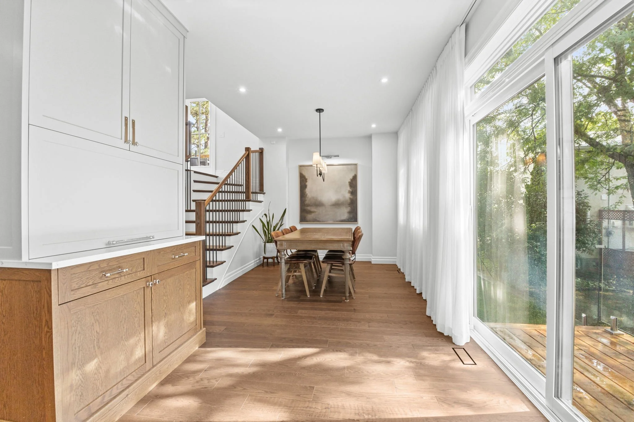 Bright dining room in a Kanata home featuring hardwood flooring, a large sliding glass door with natural light, and a wood table with seating for six, complemented by a staircase with wood railing and clean, modern finishes.