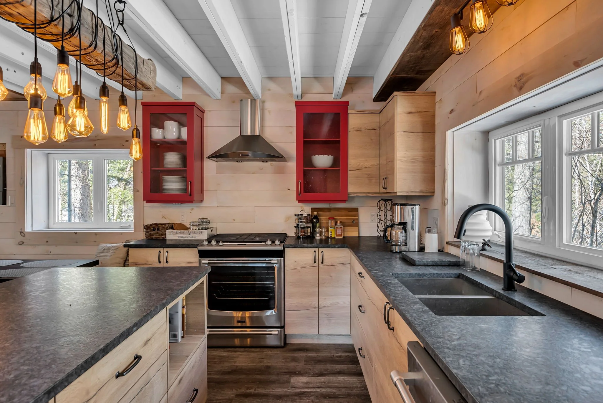 Ontario custom kitchen with wood cabinets, black countertops, stainless steel stove and range hood, black faucet, large windows, and exposed beams with Edison bulb lighting by Centennial Construction Management.