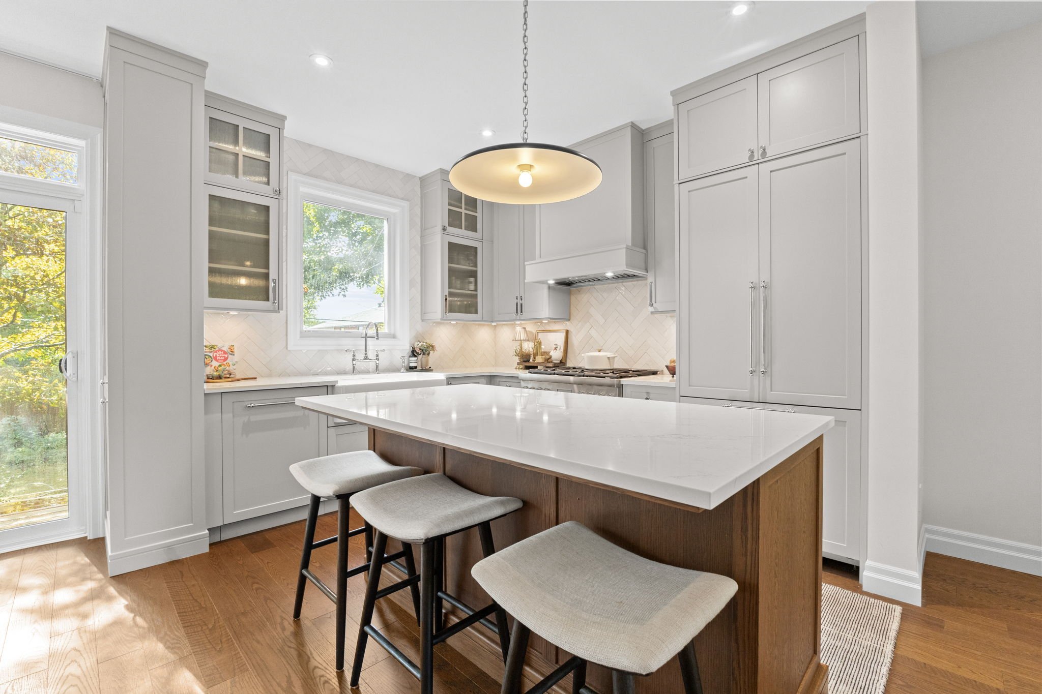 Modern kitchen with white cabinets, a large island with a white countertop, and wooden stools. There is a window above the sink, and a pendant light hanging over the island. Hardwood floors and natural light fill the space.
