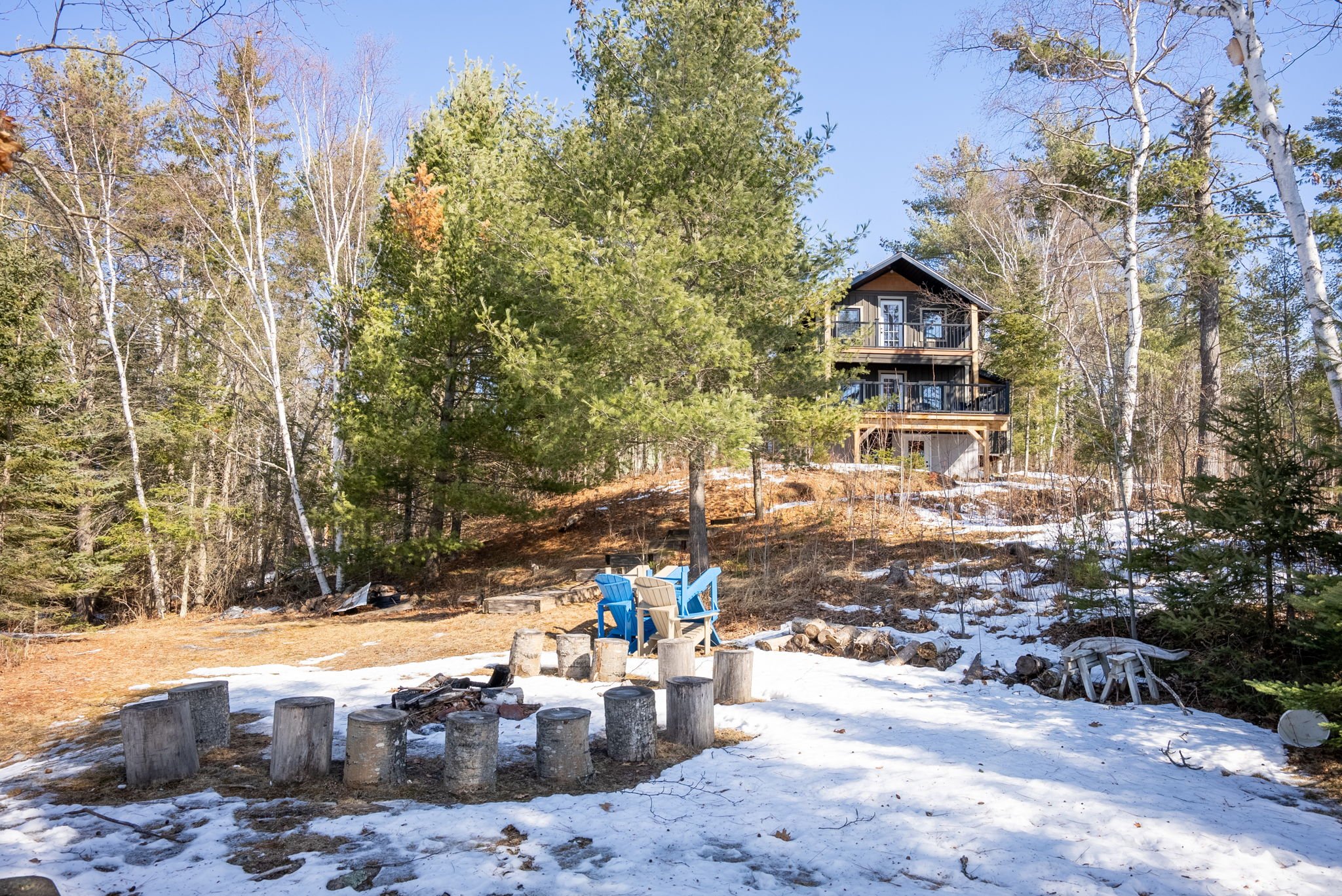 Ontario backyard landscape with fire pit circle, log stools, Adirondack chairs, and a custom home set on a wooded hillside, designed for outdoor living by Centennial Construction Management.