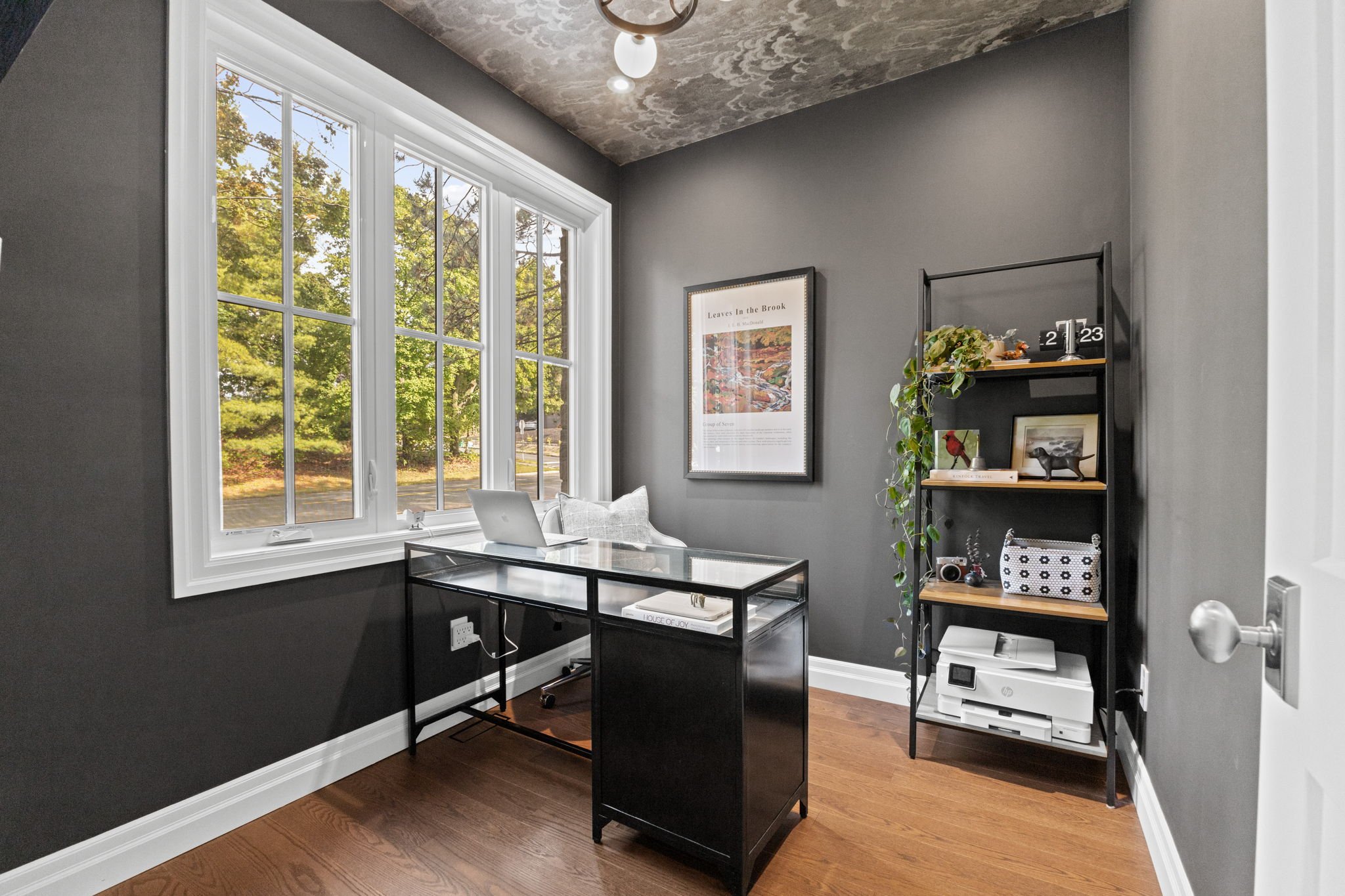 Small home office with black desk and shelving unit, large window with a view of green trees, gray walls, and wooden floor.
