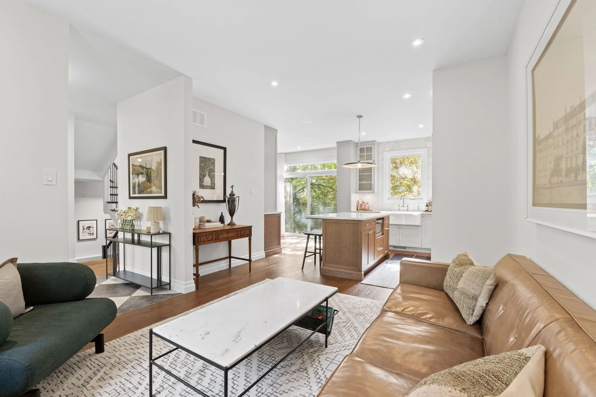 Living room with a tan leather sofa, green armchair, white marble coffee table, and wooden floor. Open kitchen in the background with a breakfast bar and window with trees outside.
