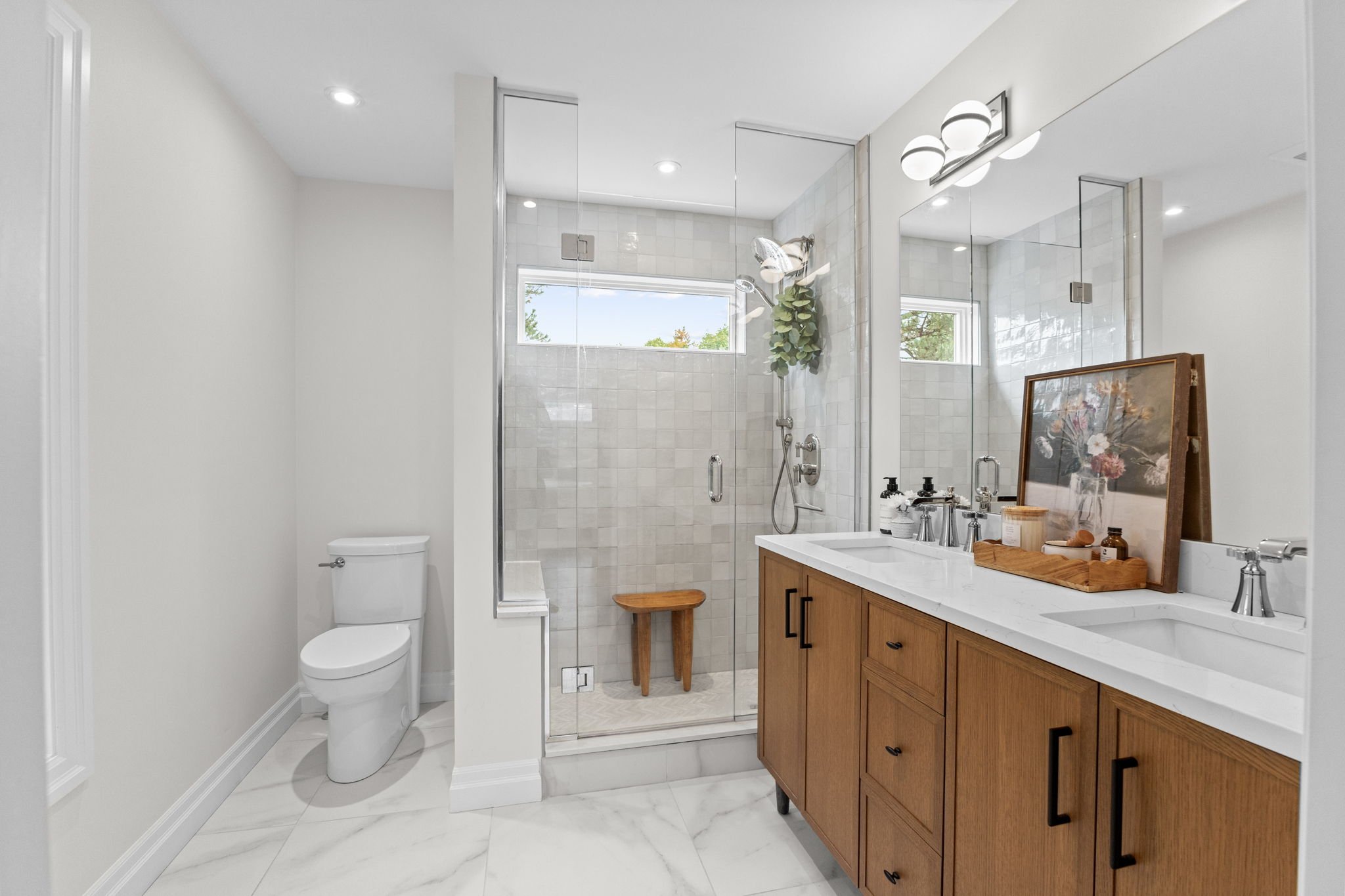 Modern bathroom with a white toilet, glass shower enclosure, wooden vanity with white countertop, mirror, and decorative items.