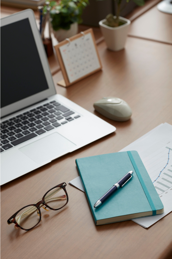 Desk with an open laptop, glasses, a closed notebook with a pen on top, a calendar, a computer mouse, a potted plant, and some papers with a graph.