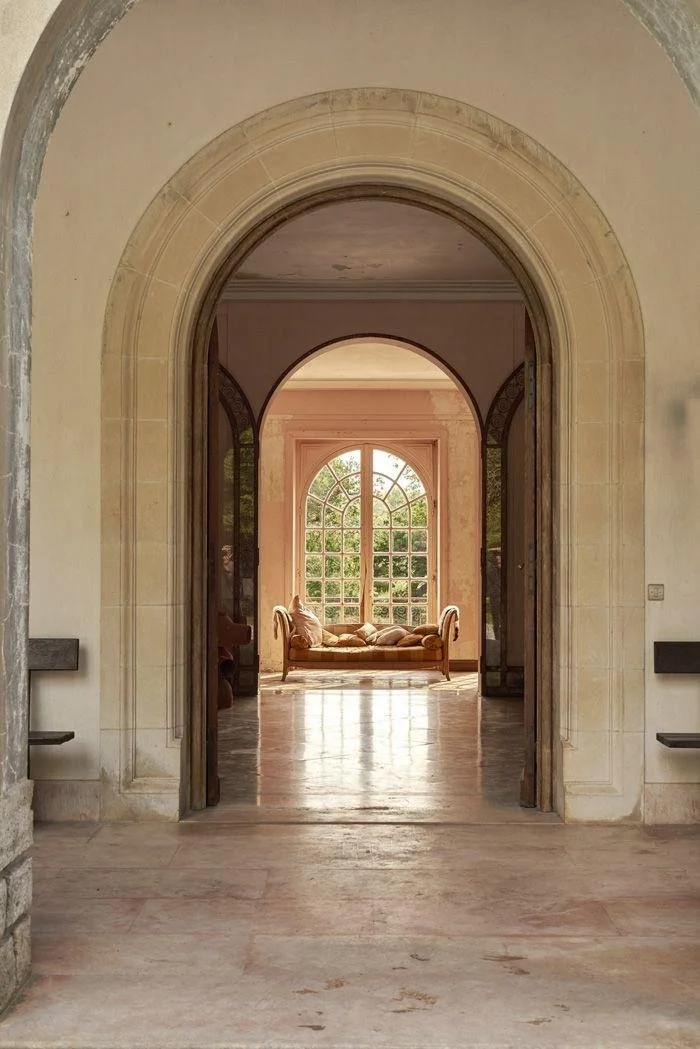 View of a spacious indoor area with multiple arches leading to a sunlit sitting room with large window and a vintage sofa.