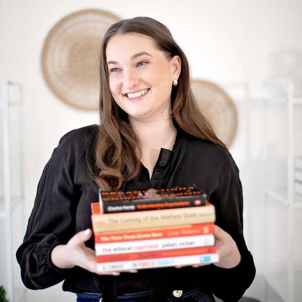 Professional photo of Rachael, she is wearing a long sleeve black adaptive shirt. She is standing and holding a stack of disability related books. She is smiling widely and looking off camera laughing.