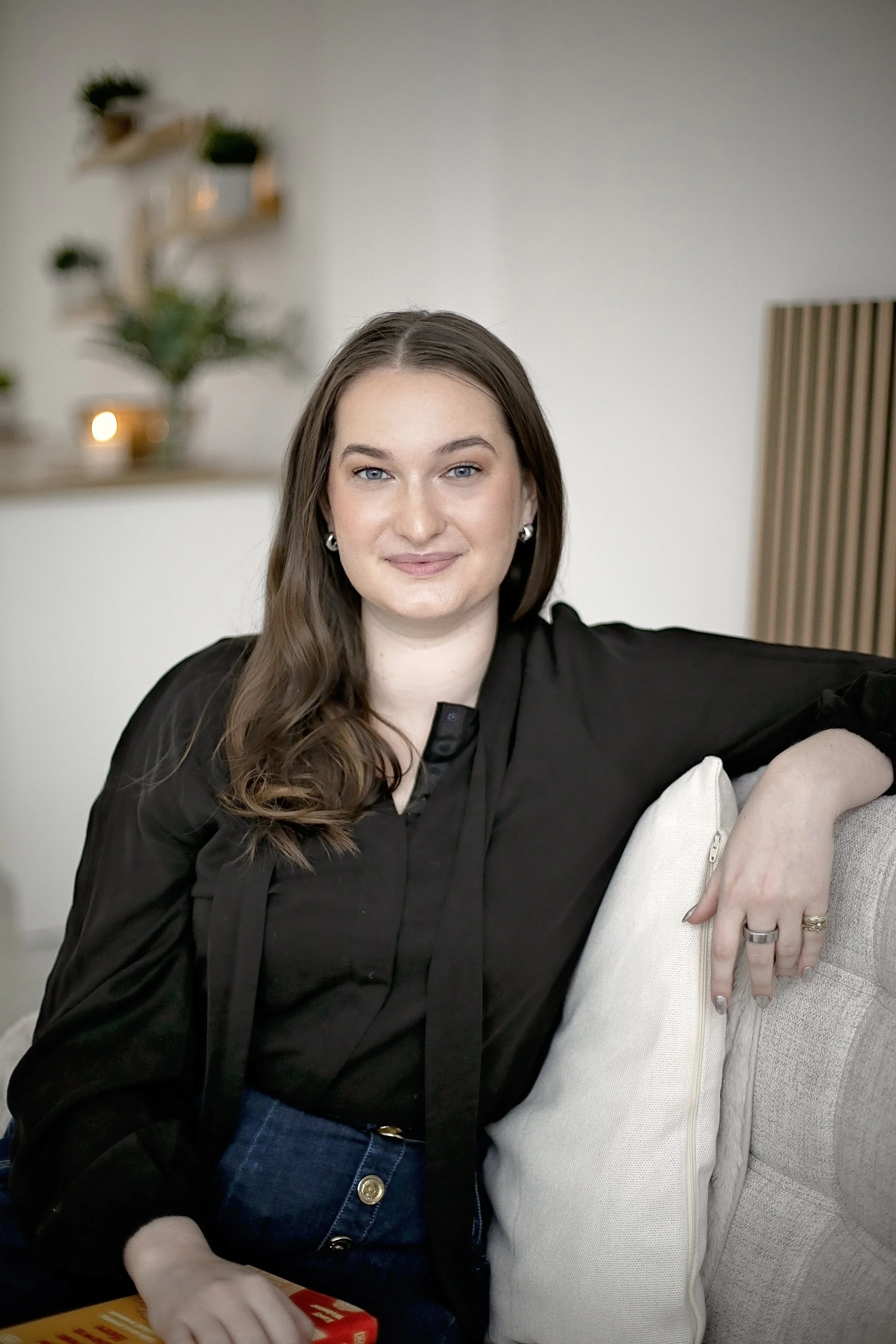 Professional headshot of Rachael, a white woman with long brown hair. She is wearing a black adaptive long sleeve top. She is sat down and has her arm over the back of the sofa.