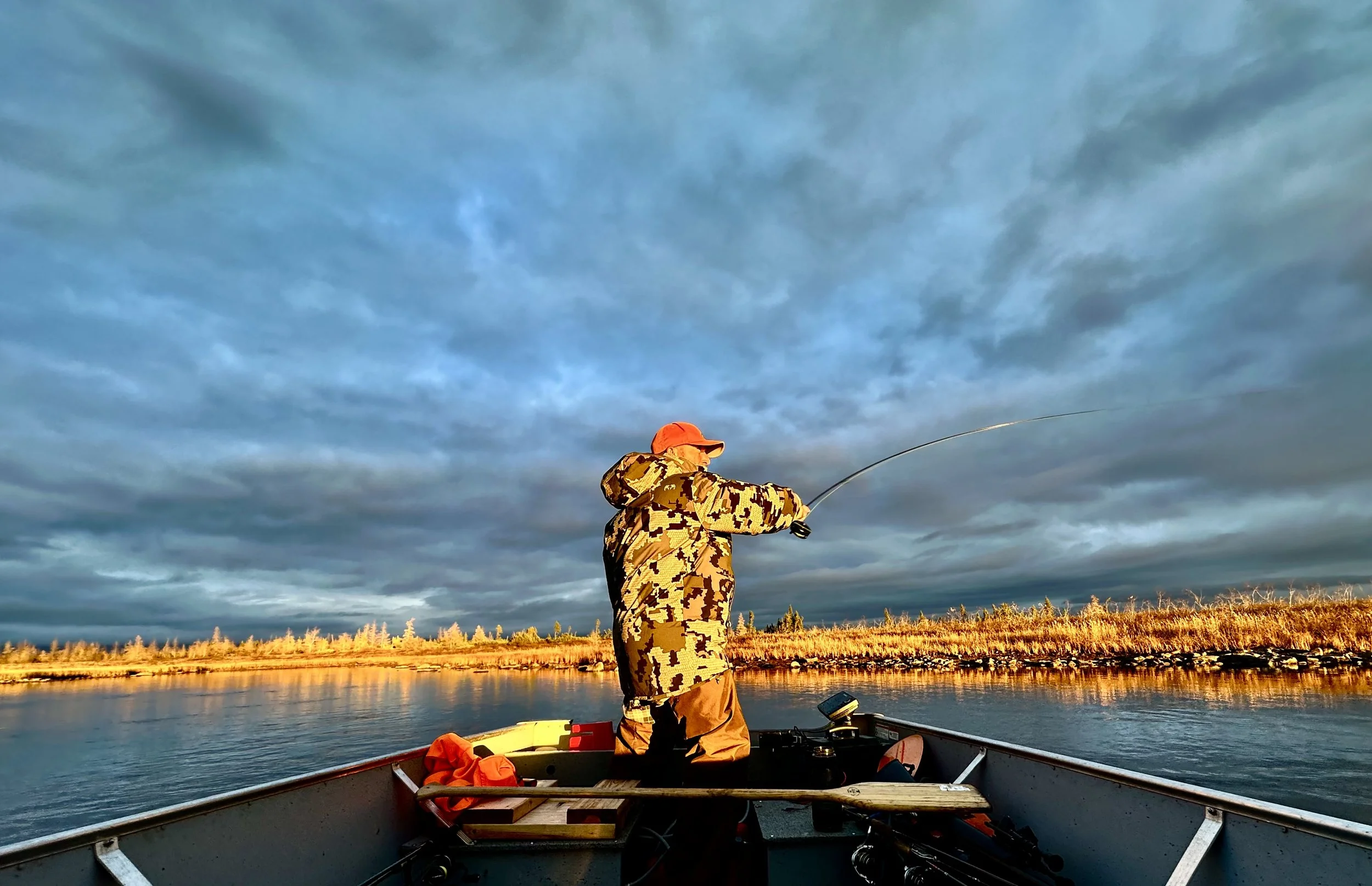 A man in camo is standing in a boat, fishing on a river, with an open tundra landscape and a cloudy sky at sunset.