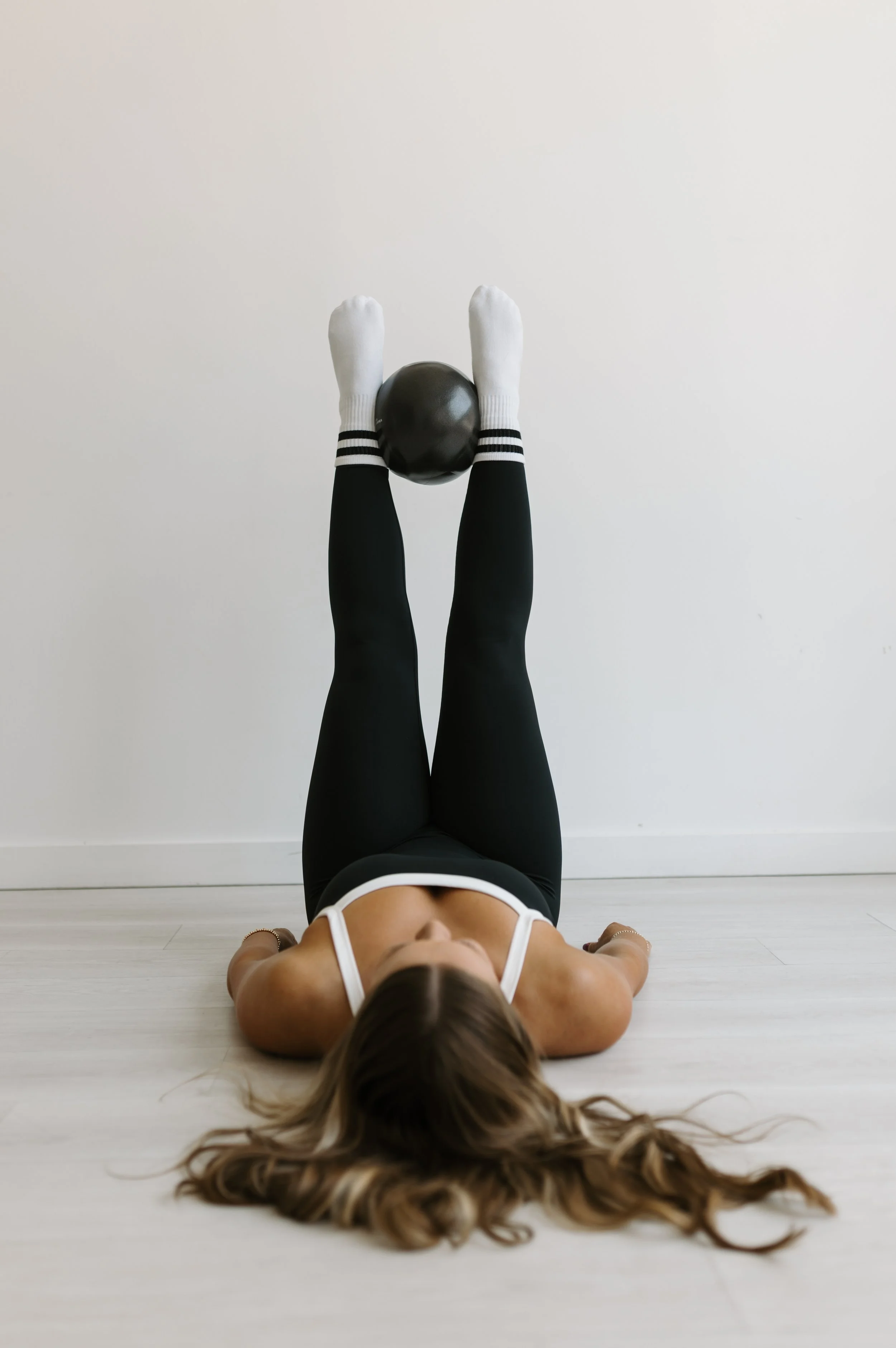 Person wearing white socks with black stripes and black pants, holding a black exercise ball straight in the air at 90 degrees while laying supine at  Worth It: A Reformer Pilates Studio in Keller, TX, near Dallas, TX & Fort Worth, Texas.