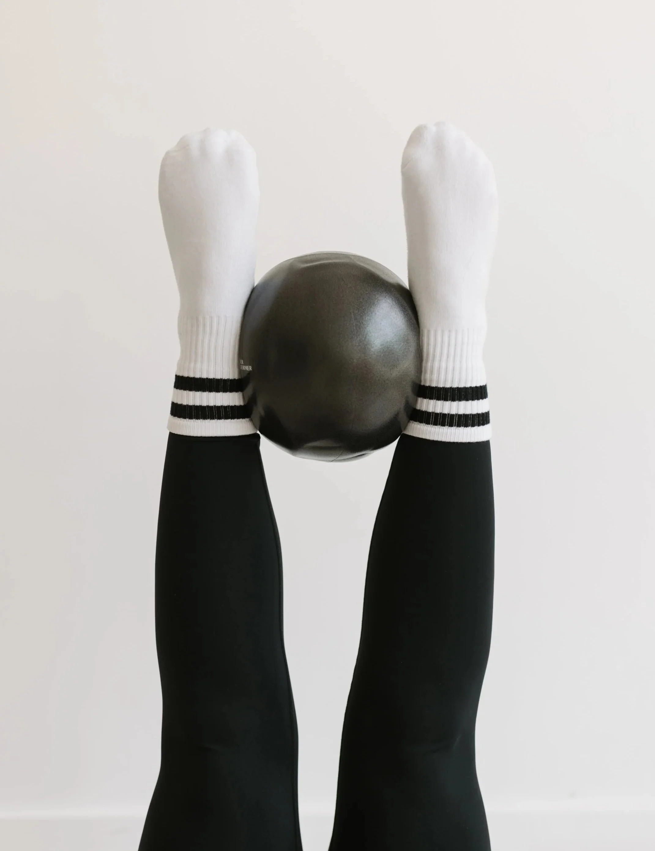 Person wearing white socks with black stripes and black pants, holding a black exercise ball straight in the air at 90 degrees on a pilates reformer at  Worth It: A Reformer Pilates Studio in Keller, TX, near Dallas, TX & Fort Worth, Texas.