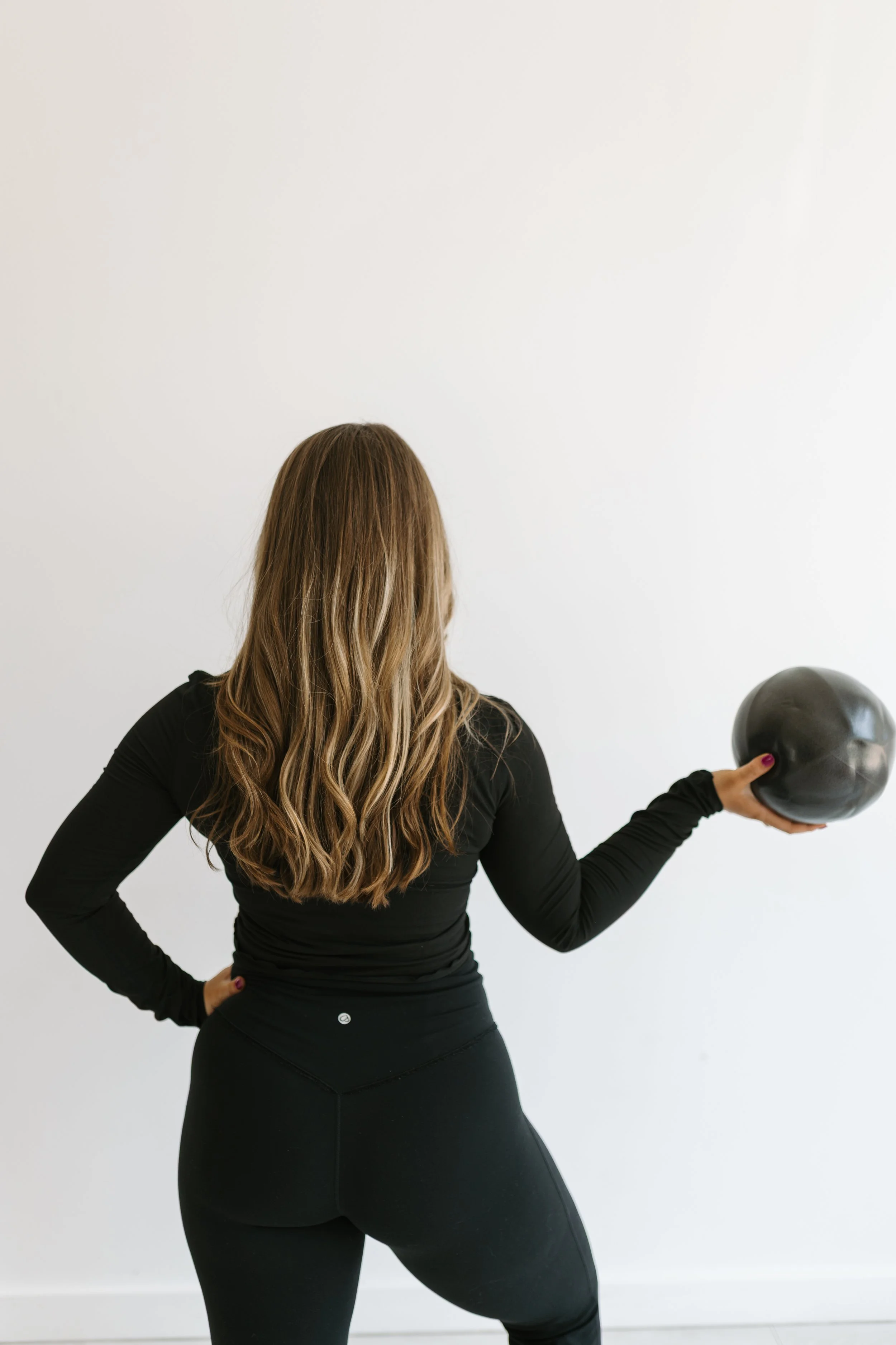Photograph of a person with long blonde hair and black workout clothes holding an exercise ball that is used during the group reformer pilates classes at Worth It: A Reformer Pilates Studio located in Keller Texas, near Dallas, Texas and Fort Worth, 