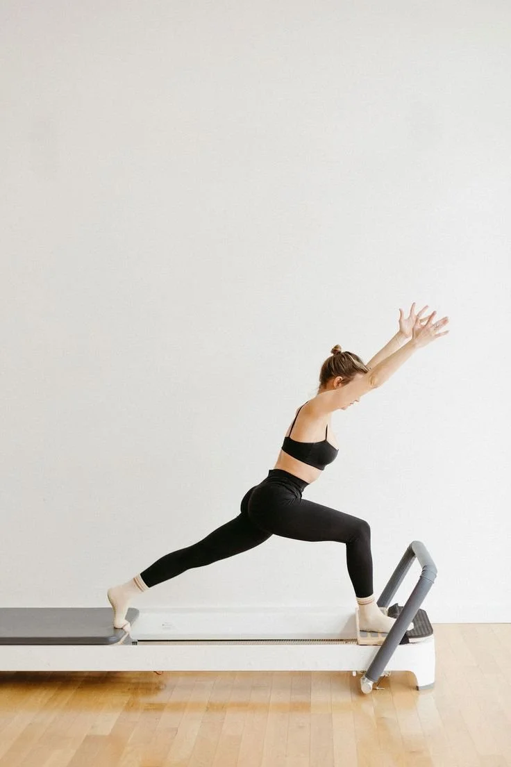 A woman in a black workout outfit performing a pilates pose on a Studio X Pilates reformer machine, with her body in a lunge with her arms raised straight above her head on the pilates reformer at Worth It: A Reformer Pilates Studio located in Keller