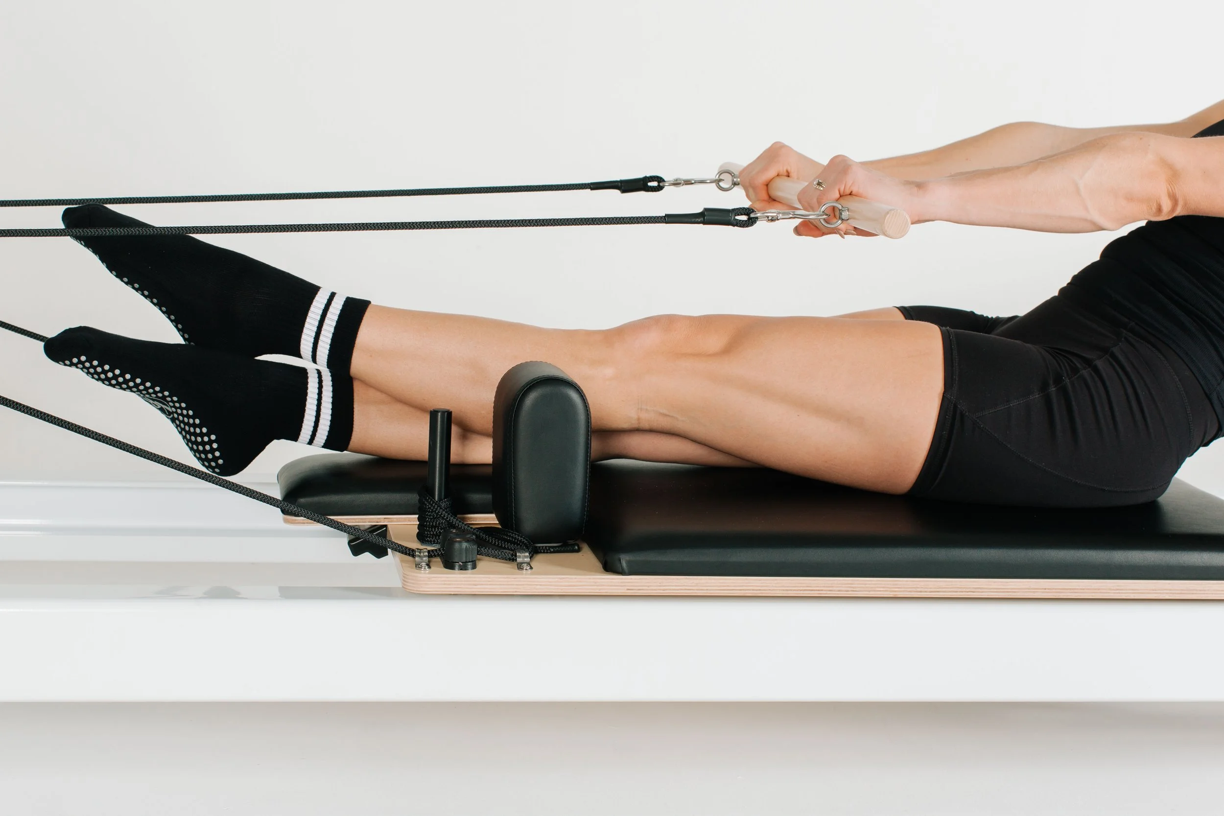 A person performing Pilates exercise on a reformer machine, lying on their back with legs extended and gripping handles attached to resistance cords.
