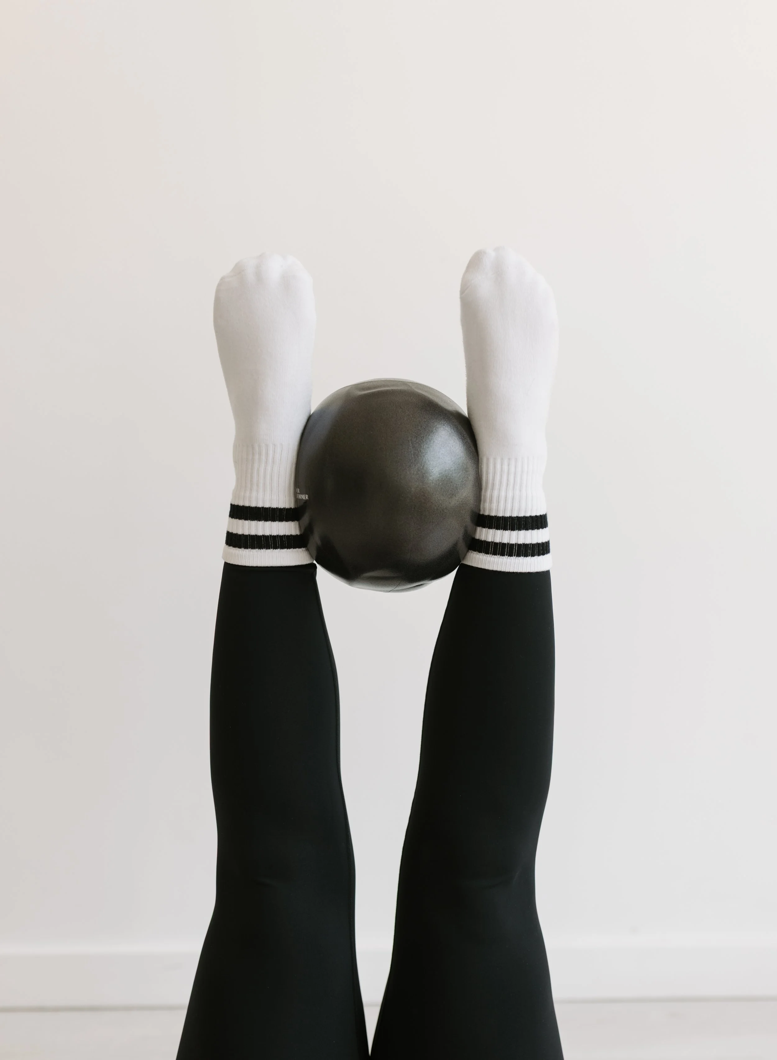 Person wearing white socks with black stripes and black pants, holding a black exercise ball straight in the air at 90 degrees on a pilates reformer at  Worth It: A Reformer Pilates Studio in Keller, TX, near Dallas, TX & Fort Worth, Texas.