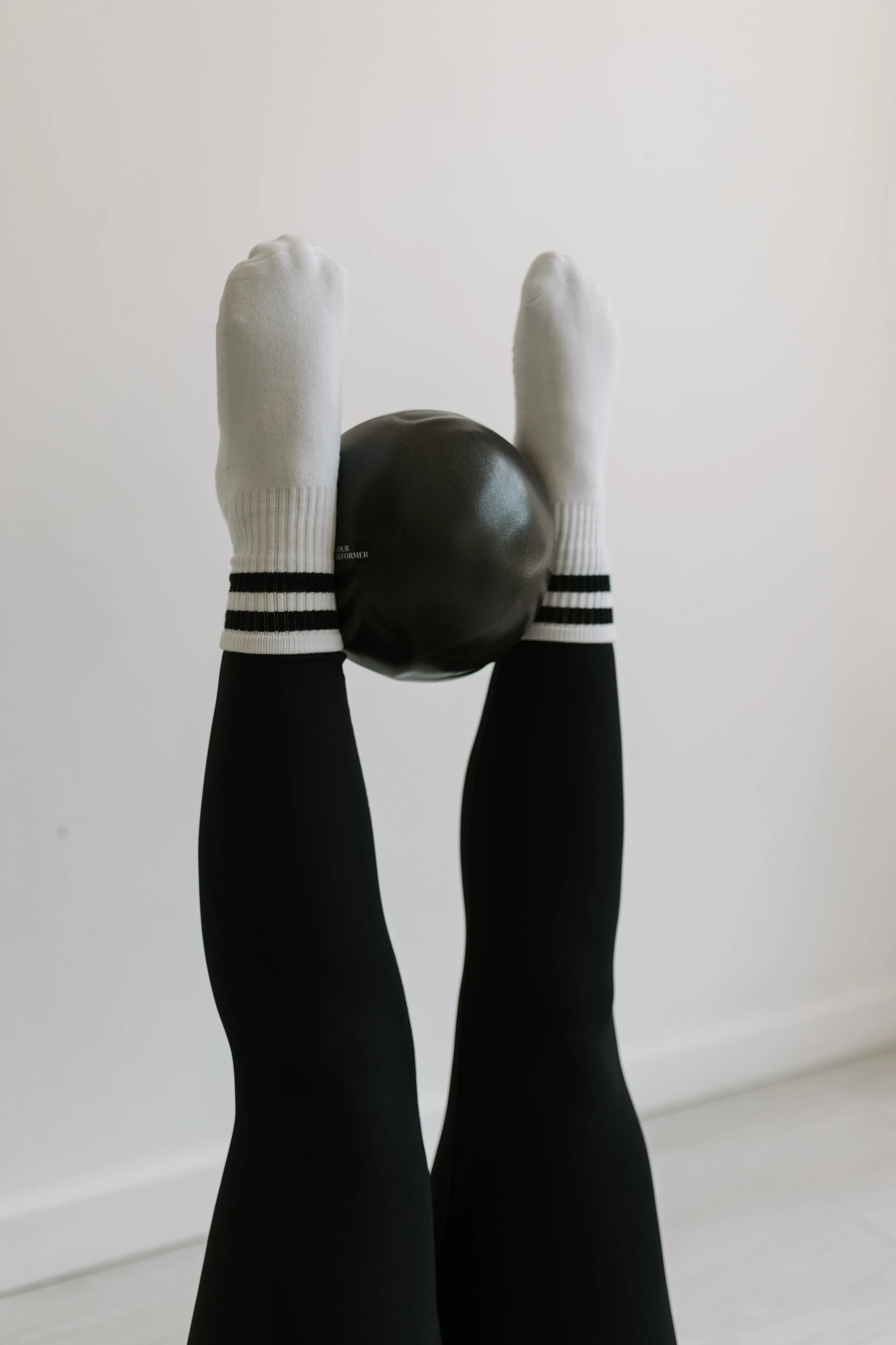 Person wearing white socks with black stripes and black pants, holding a black exercise ball straight in the air at 90 degrees on a pilates reformer at  Worth It: A Reformer Pilates Studio in Keller, TX, near Dallas, TX & Fort Worth, Texas.