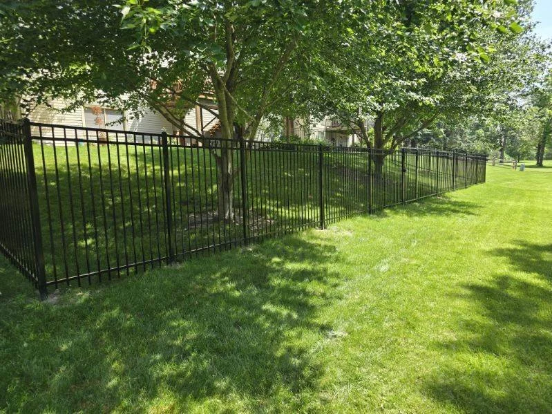 Black metal fence running along a grassy yard with trees and a house in the background.
