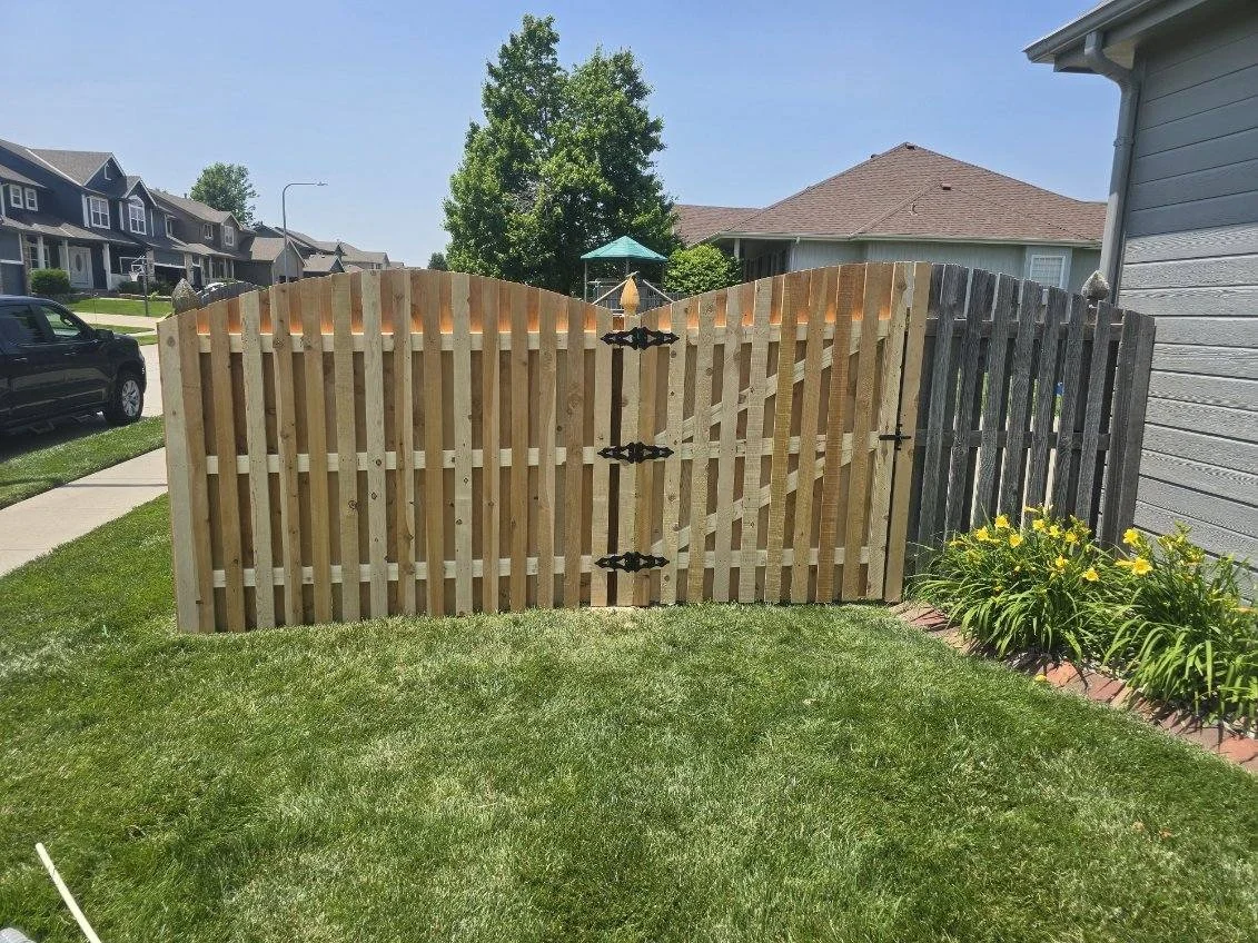 A wooden backyard gate with a curved top, connected to a fence, with green grass and yellow flowers nearby, houses and trees in the background under a clear sky.