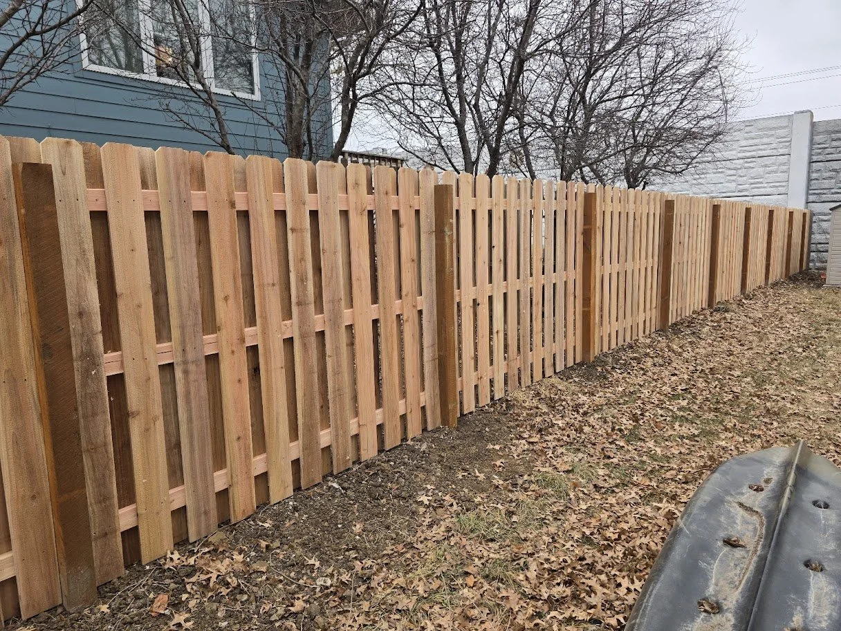 New wooden fence installed in a backyard with fallen leaves on the ground. A blue house with windows and leafless trees are in the background.