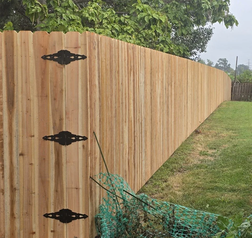 A long wooden privacy fence with decorative black hinges, with greenery and trees in the background, and a patch of grass in the foreground.