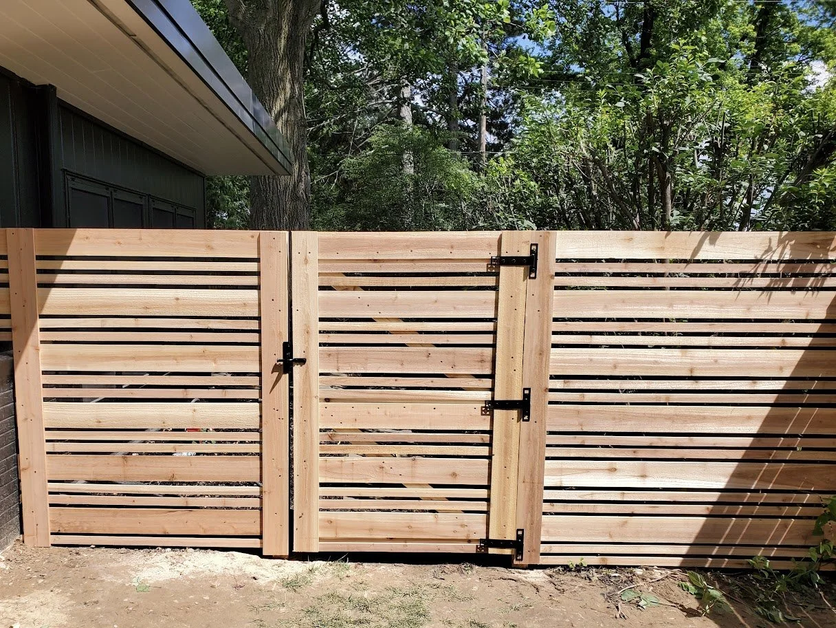 A new wooden fence with a gate, installed in front of a house and surrounded by trees and greenery.