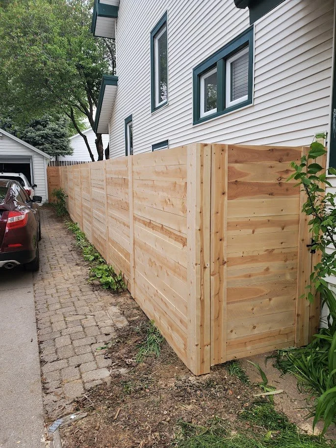 New wooden privacy fence installed along the side of a house with white siding and green window frames, next to a driveway with a parked car, in a residential area with trees.