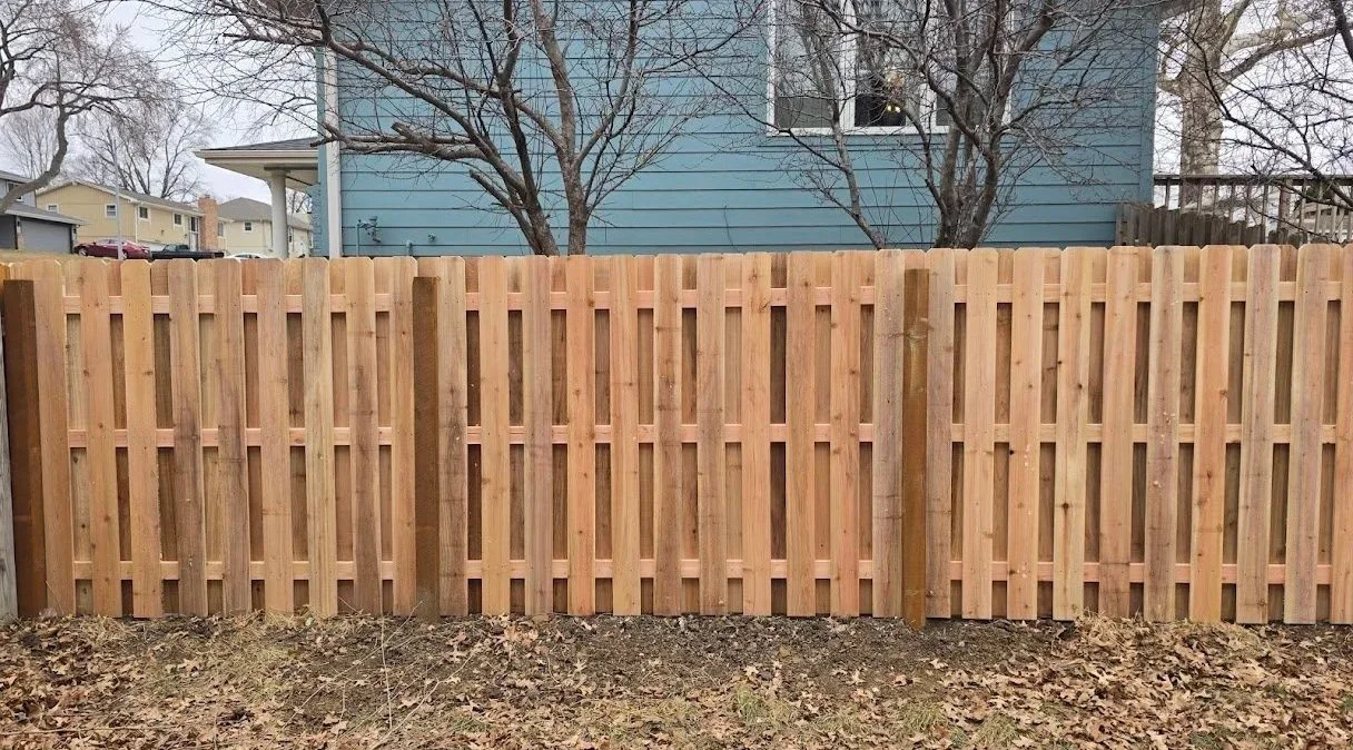 New wooden privacy fence in a backyard with trees and a blue house in the background.