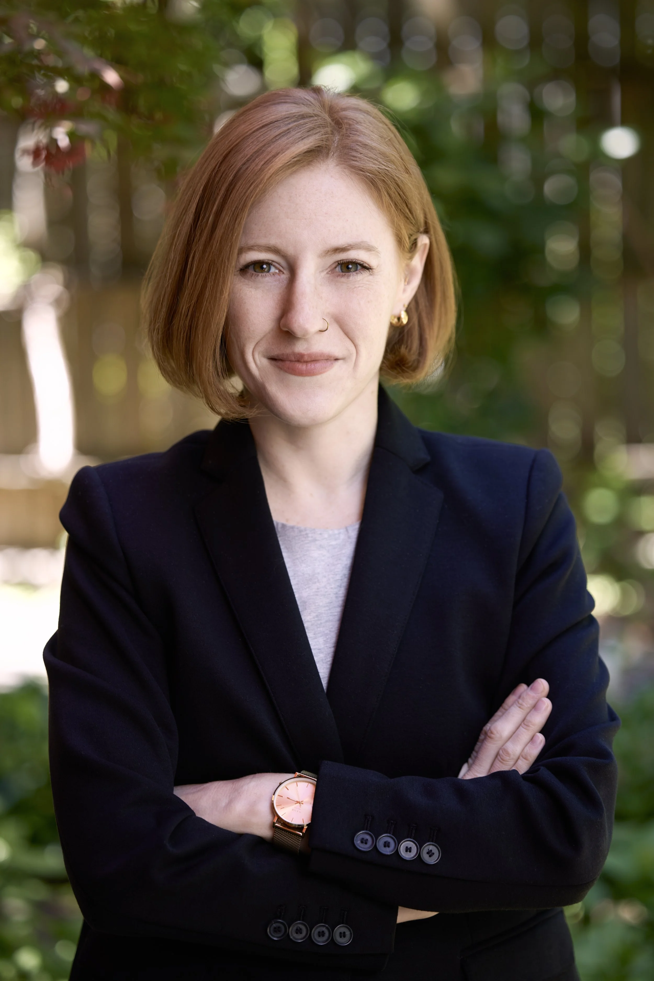 A woman with red hair and light skin wearing a black blazer, light gray shirt, gold earrings, a small nose ring, and a gold watch with a pink face, standing outdoors with green blurred trees in the background, arms crossed.