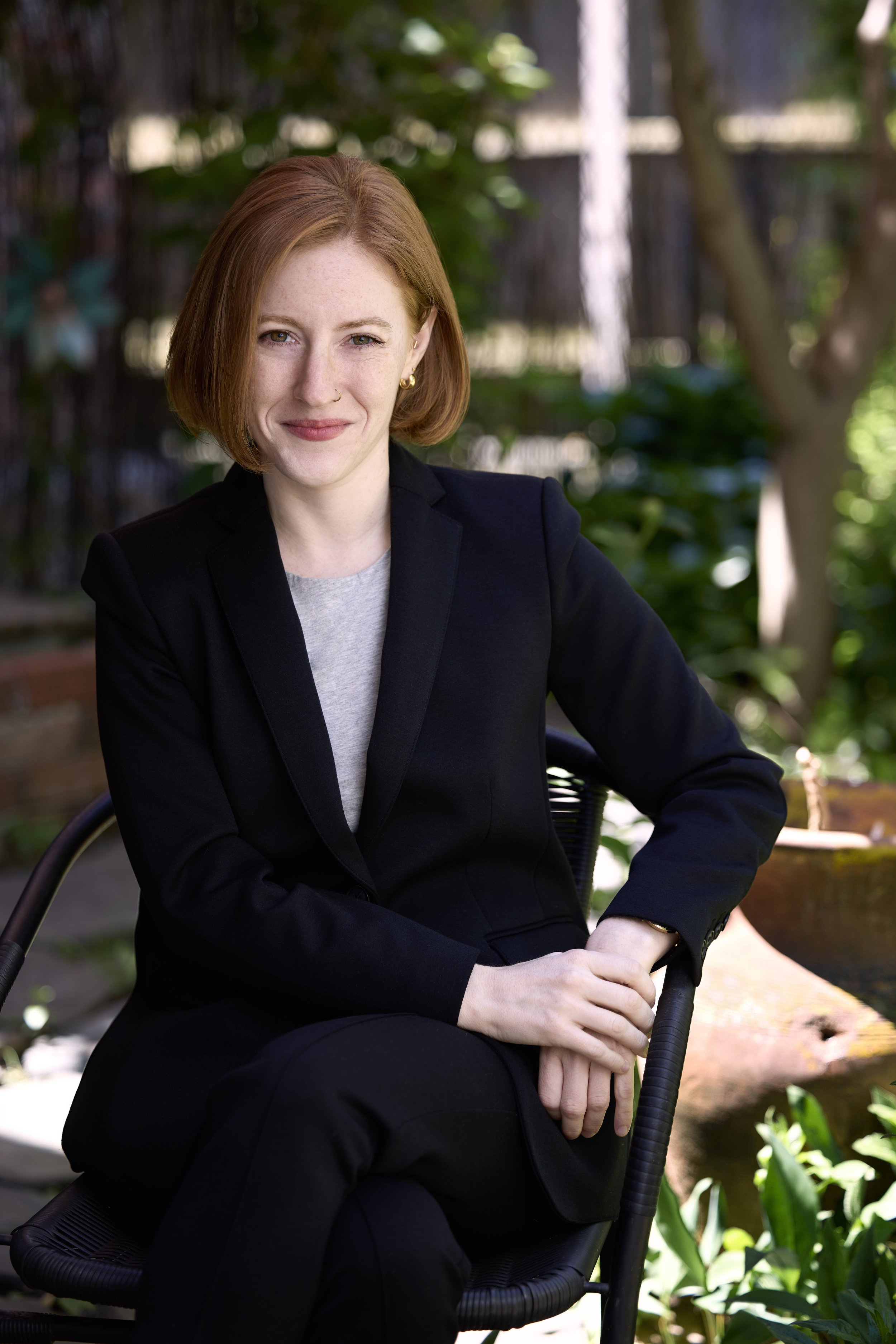 A woman with red hair, wearing a black blazer and gray shirt, sitting outdoors in a garden setting with trees and greenery in the background.