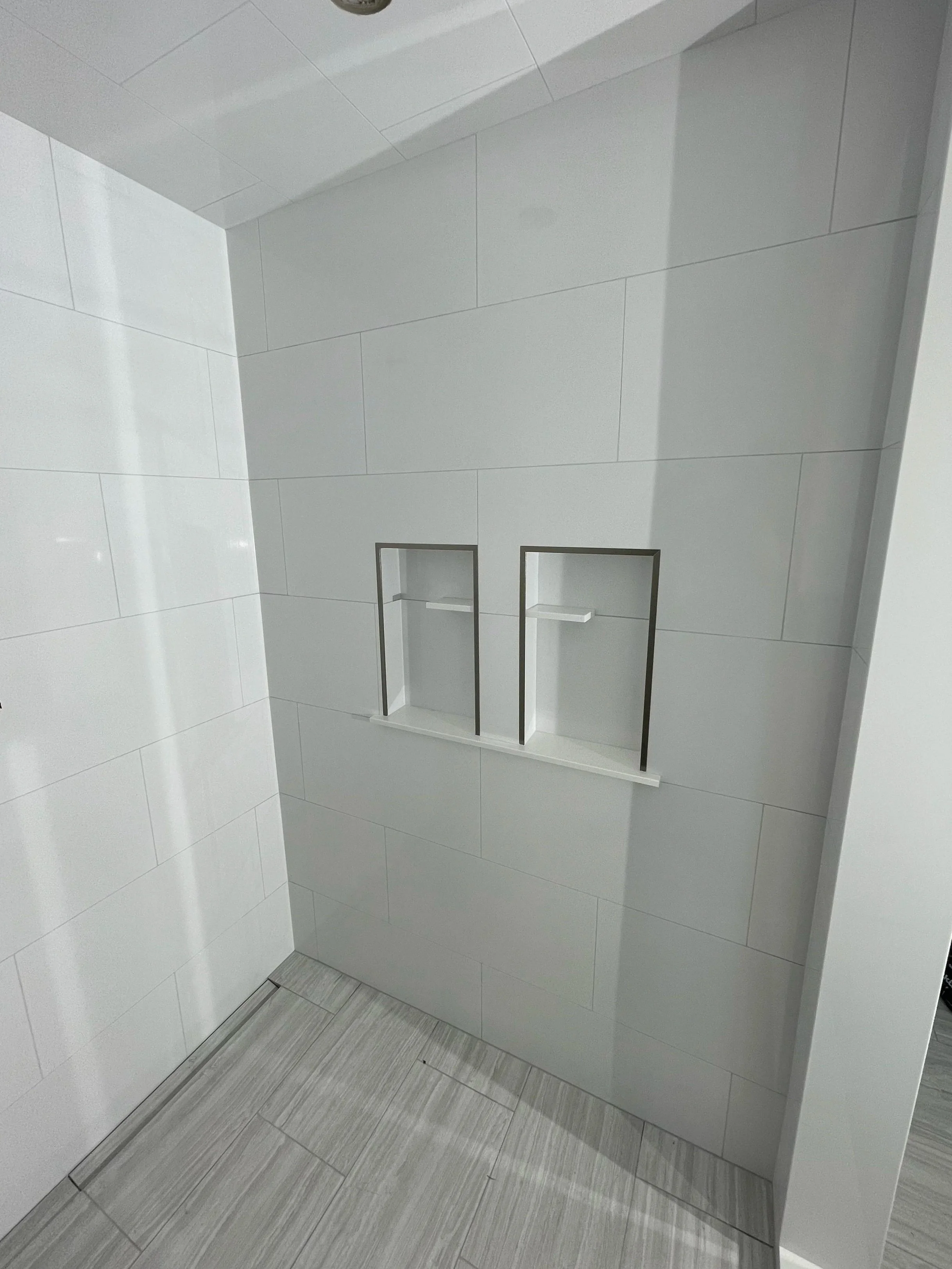 A modern shower wall with two built-in recessed shelves, tiled in large white tiles, with a light-colored tiled floor.  Tile Work and Contractor Kawartha Lakes, GTA, Muskokas. Custom tile work with backlighting. Bathroom renovation. 