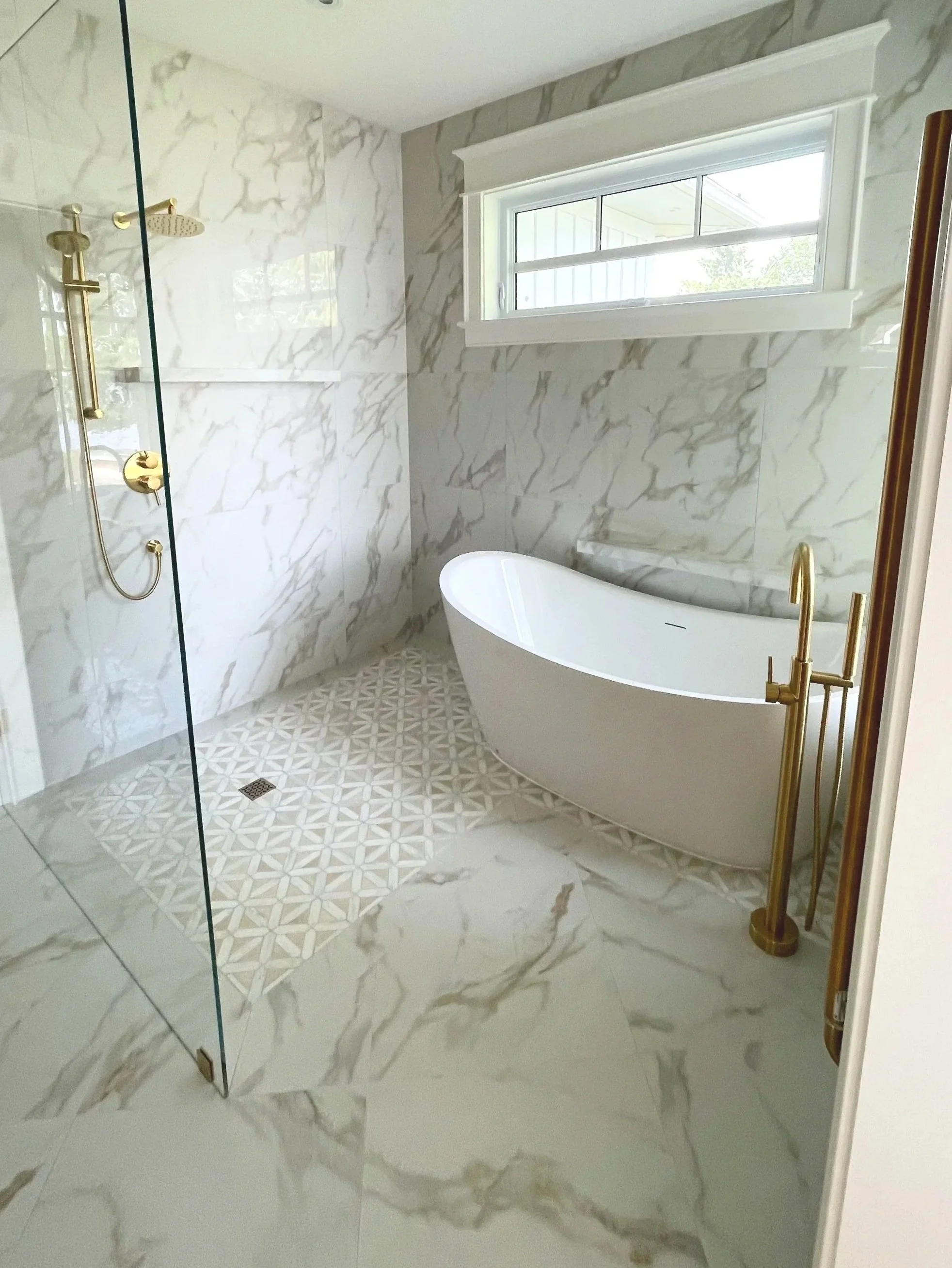 A modern bathroom with a freestanding bathtub, marble walls and floor, a window above the bathtub, and a glass shower enclosure with gold fixtures.  Tile Work and Contractor Kawartha Lakes, GTA, Muskokas.