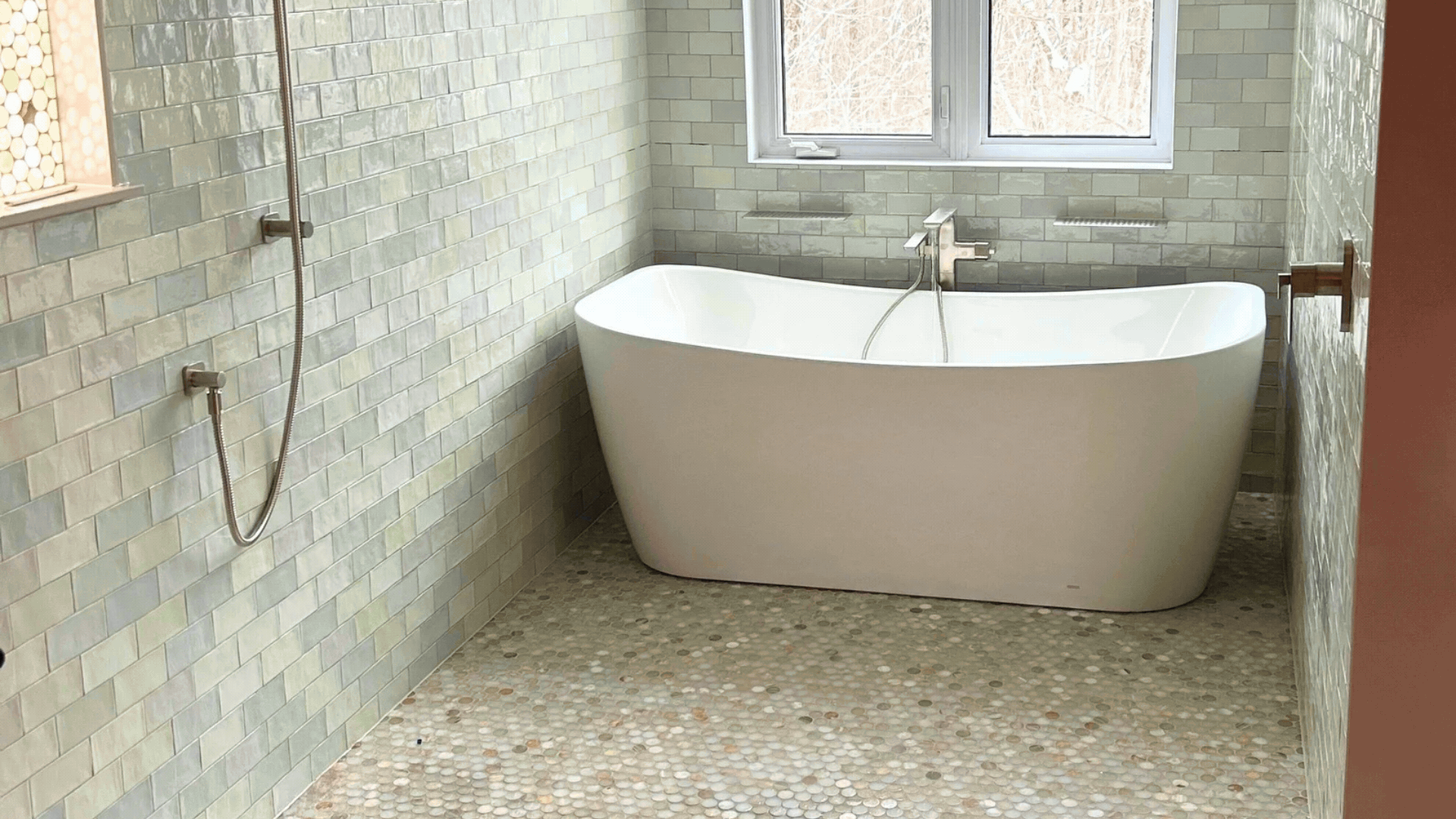 A modern bathroom featuring a freestanding white bathtub positioned under a window with tiled walls and pebble flooring.  Tile Work and Contractor Kawartha Lakes, GTA, Muskokas.